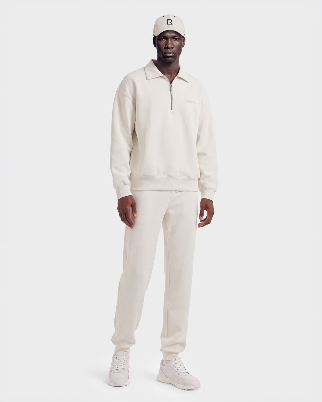 A man wearing the Rudsak IDRIS - 6526500 Vanilla zip-collar sweatshirt with matching sweatpants, sneakers, and a beanie stands against a plain white background.