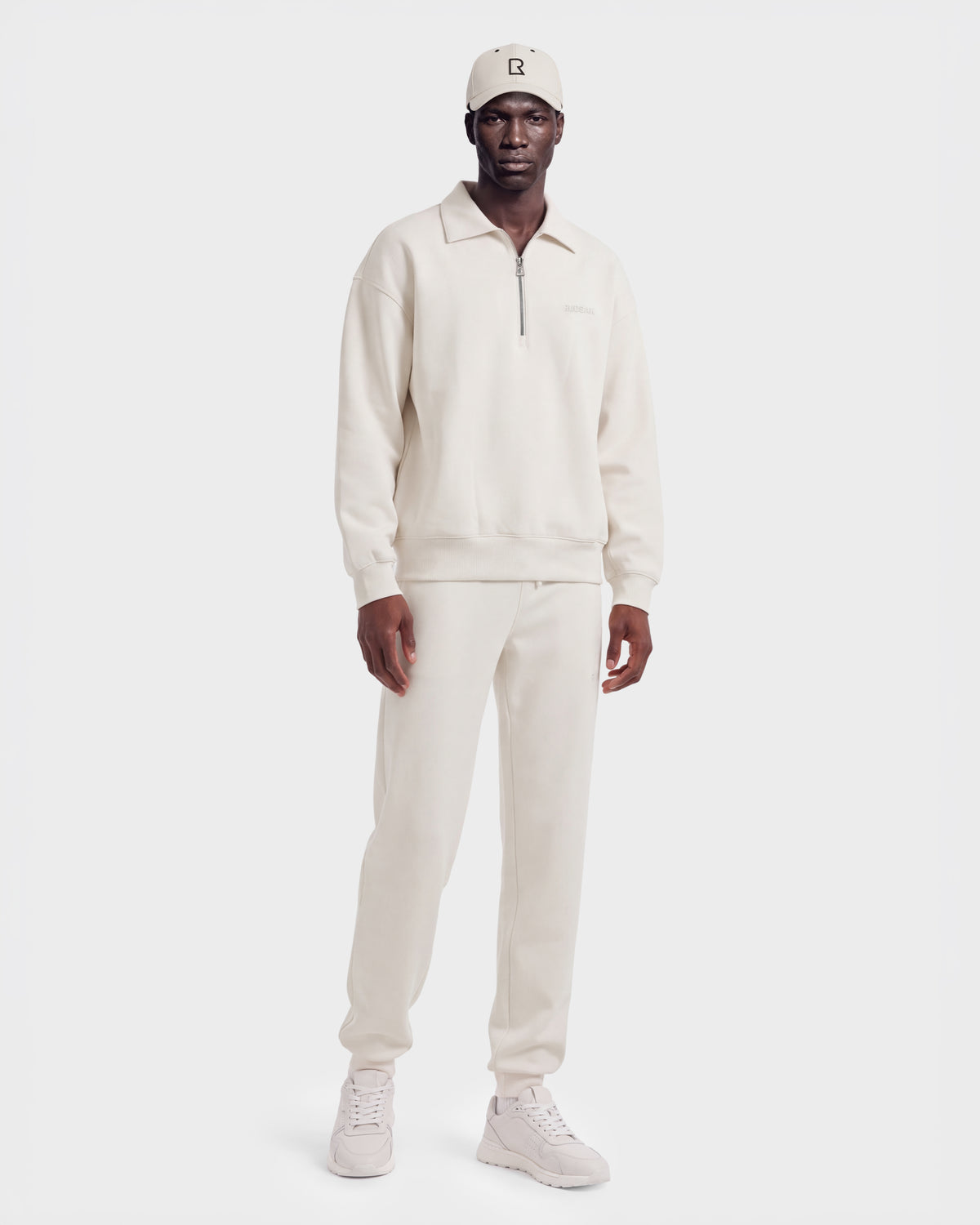 A man wearing the Rudsak IDRIS - 6526500 Vanilla zip-collar sweatshirt with matching sweatpants, sneakers, and a beanie stands against a plain white background.