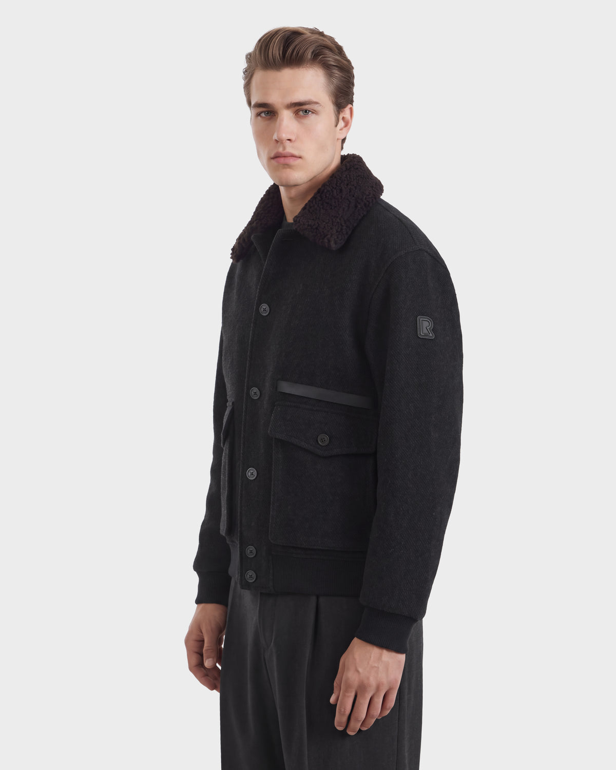A man wearing a JULIUS - 6126723 Charcoal bomber jacket in dark charcoal wool-blend with a dark faux-fur collar, flap pockets, and a ribbed hem.