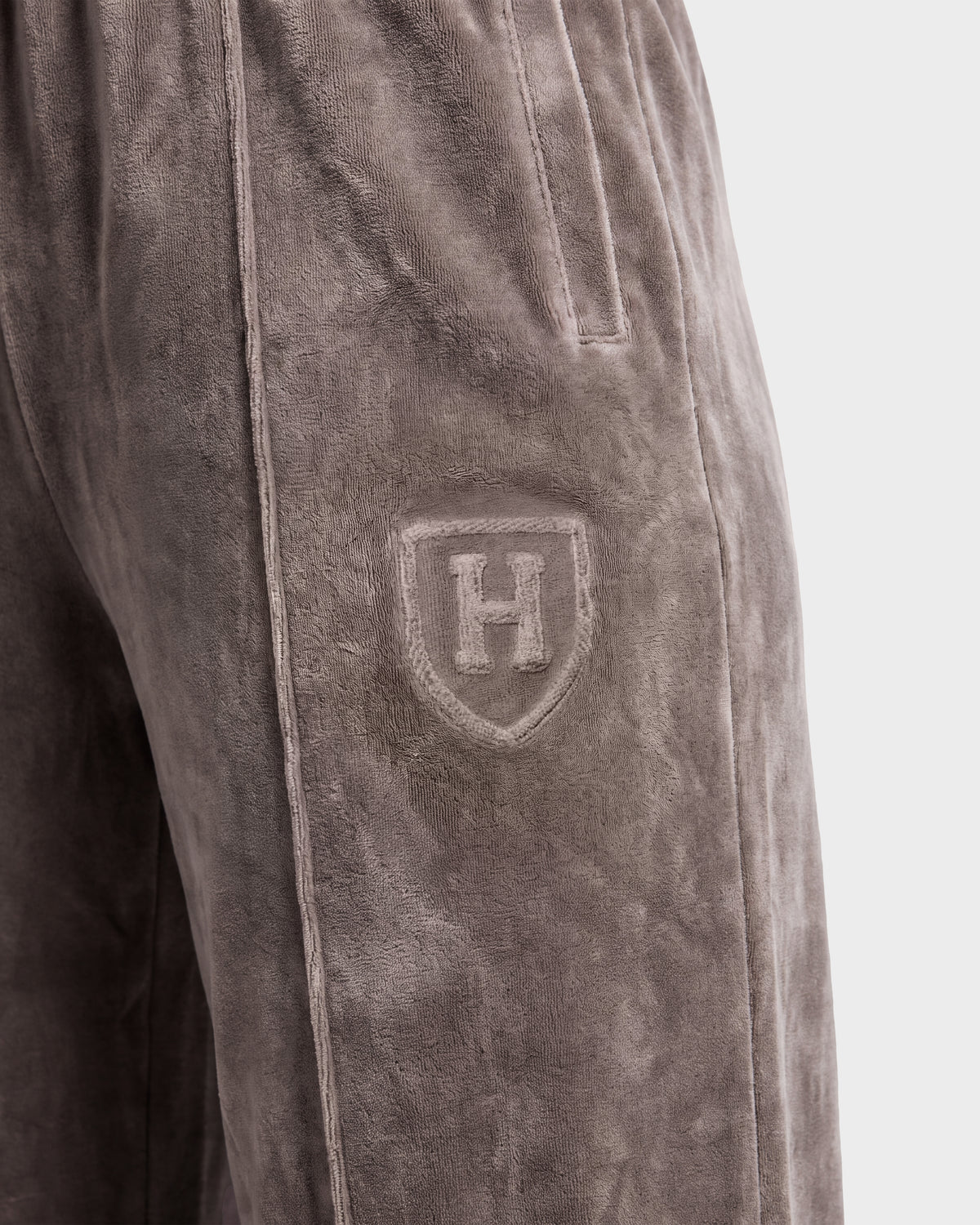Close-up of the Rudsak OLIVYA HV - H526903 Taupe velour pants, featuring a side pocket and an embroidered shield logo with the letter H on the upper thigh.