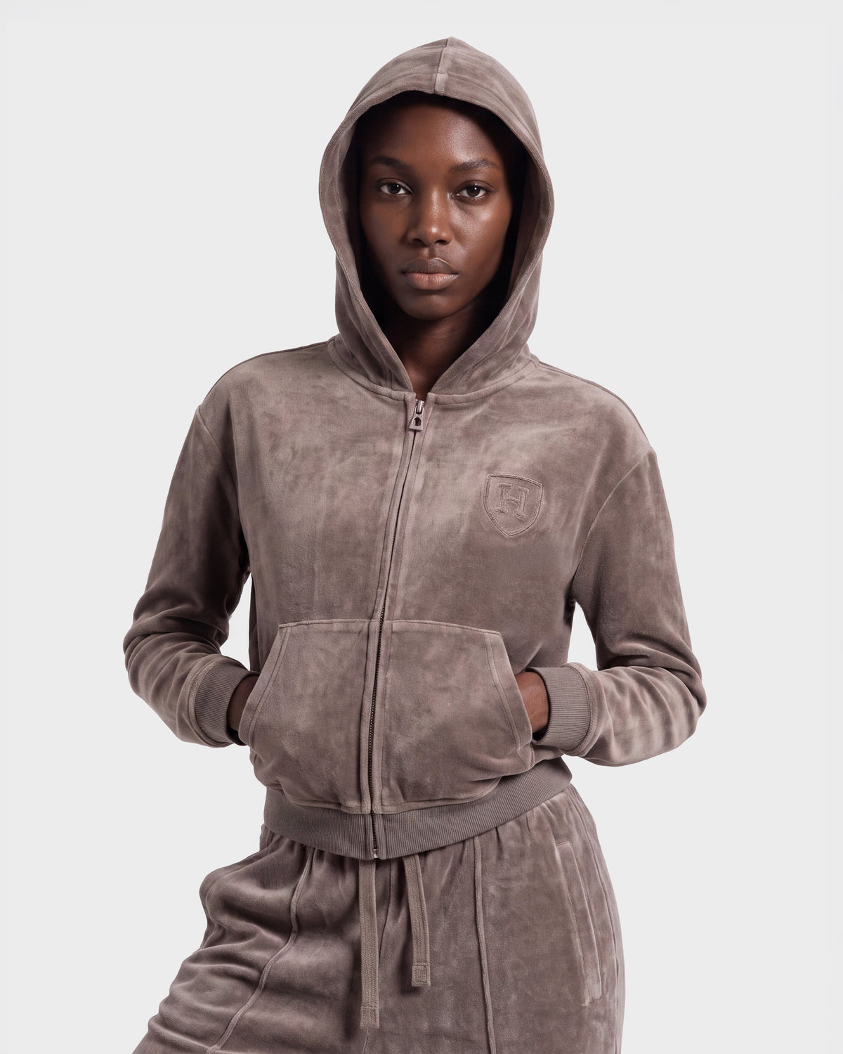 A person wearing the Rudsak TYRA HV - H526904 Taupe velour tracksuit with the hood up and hands in the front pockets, standing against a plain light background.