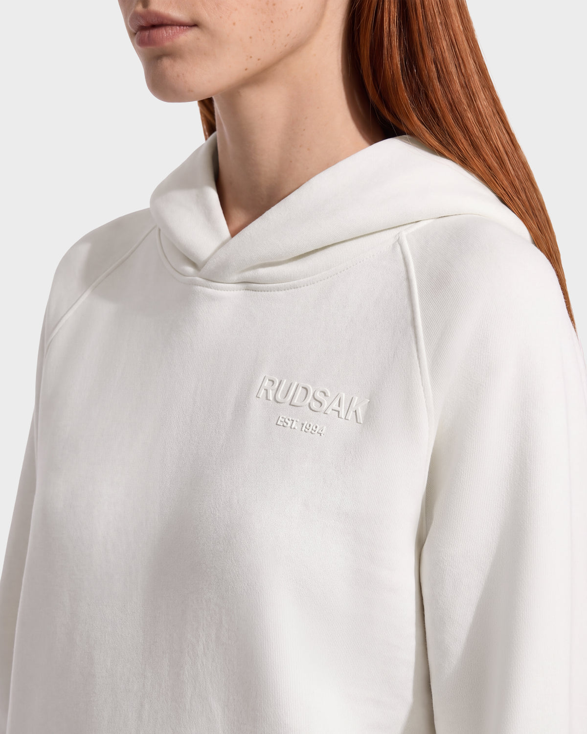 Person wearing a white BRIAR - 9525001 Vanilla W unisex hoodie featuring an embossed logo on the chest that reads RUDAK EST. 1994, completing a casual look.