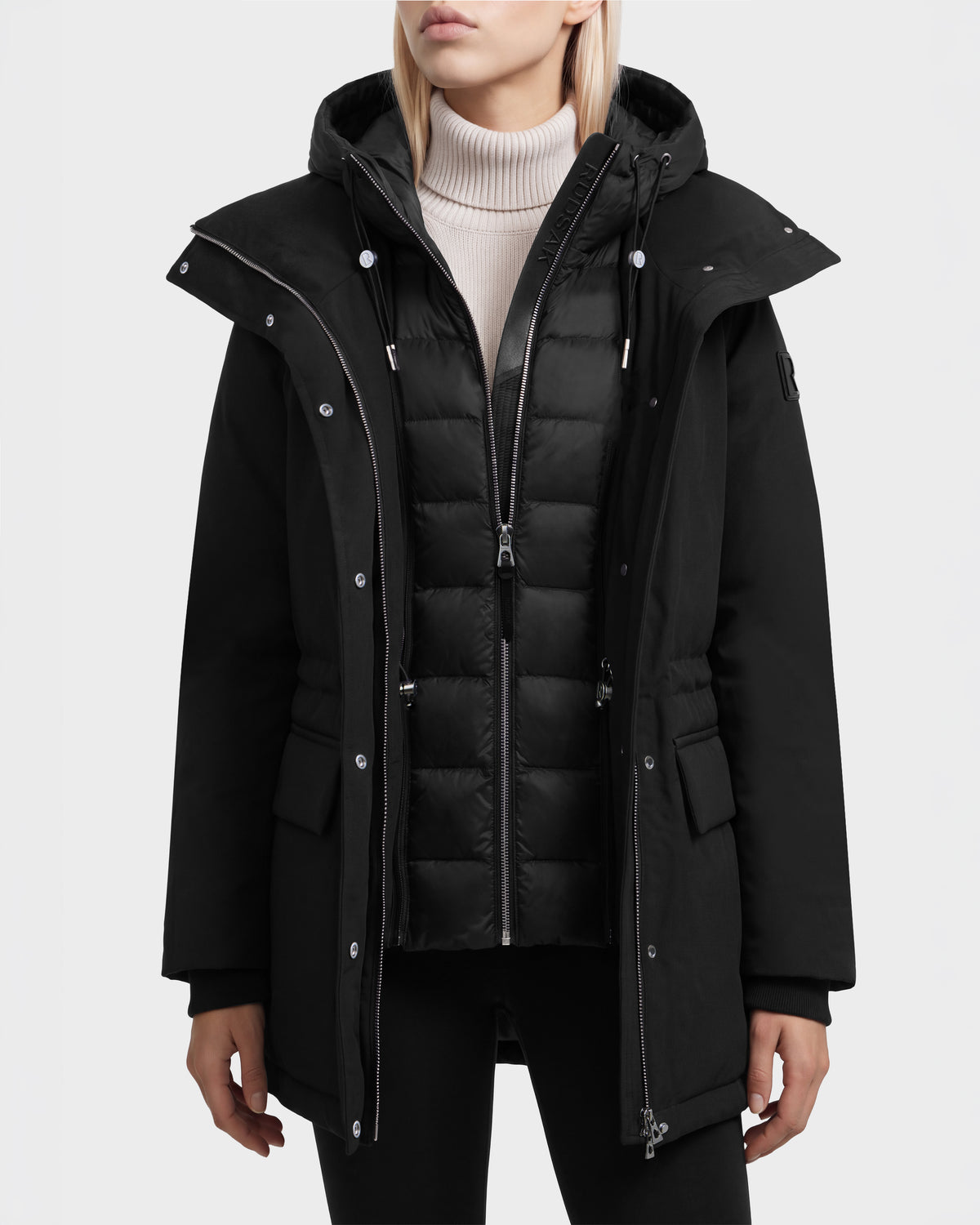 A person wearing a black Rudsak BHIMA - 8126515 Black parka featuring an integrated quilted vest, a hood, and a zippered front.