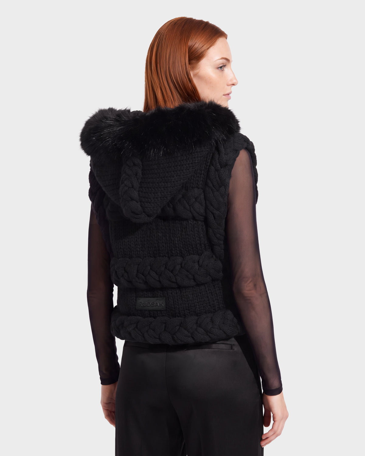 A woman with red hair, seen from behind against a plain background, models the Rudsak CICELY - 8526686 Black sleeveless knit vest featuring a faux fur-trimmed hood—ideal for winter layering—styled over a sheer black top and black pants.