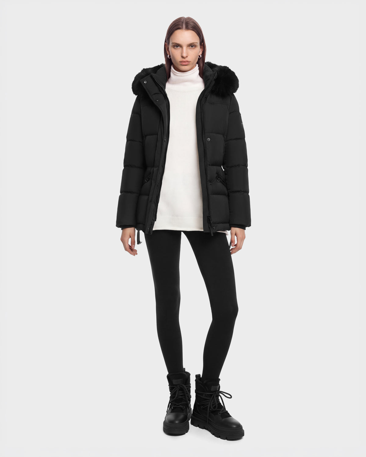Model wearing a Rudsak GRACE FF - 8126535 Black hooded puffer jacket with fur trim over a white turtleneck, black leggings, and black lace-up boots.