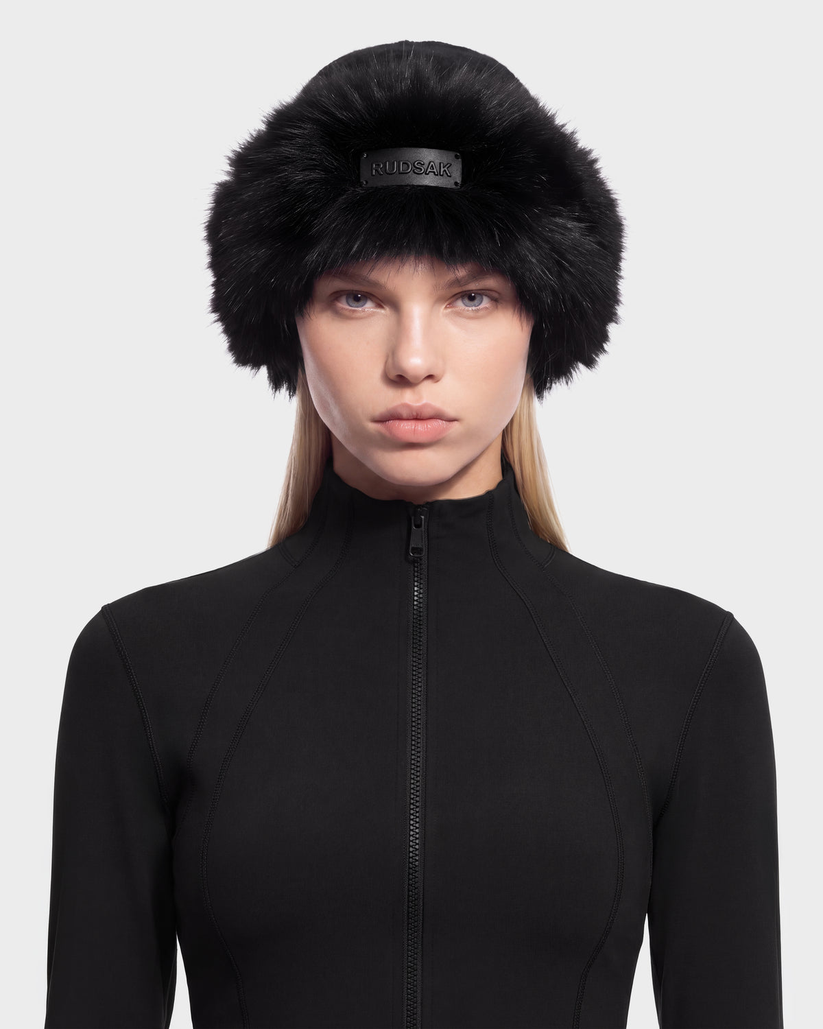 A person wearing a black faux fur LORETTA W - 8426510 Black cossack-style hat with a logo plaque on the front and a black zippered mock-neck top.