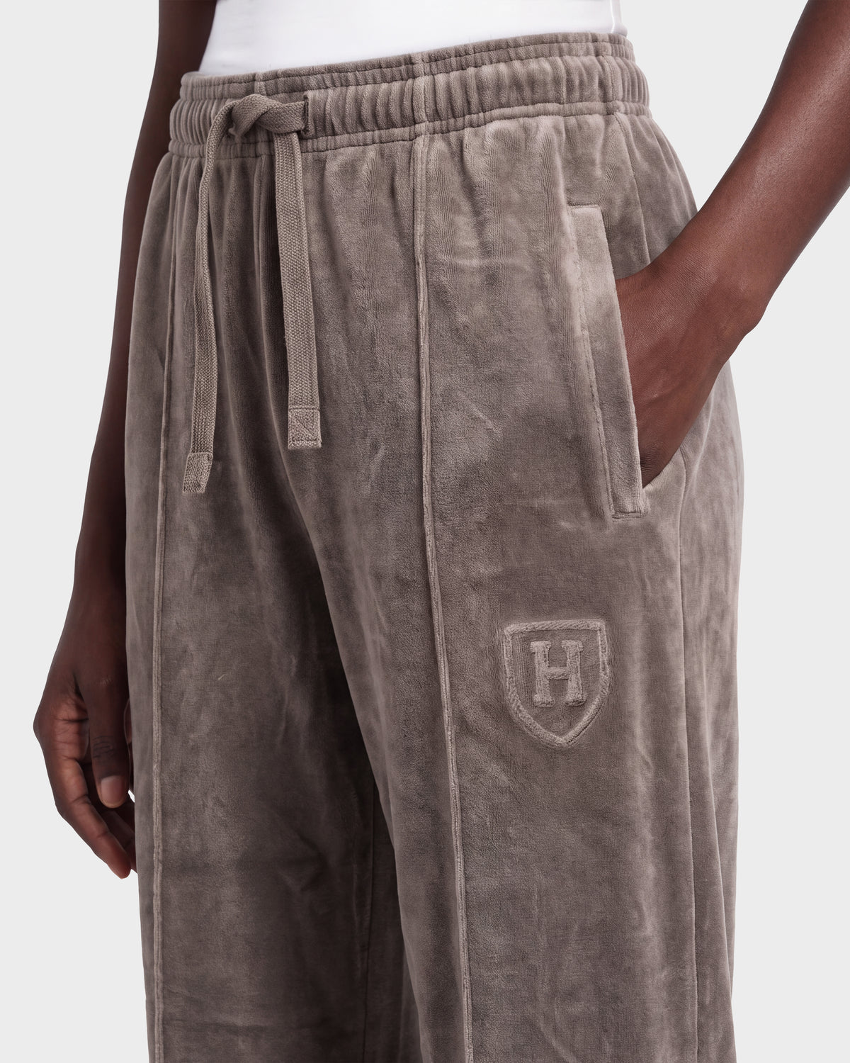 A person wearing Rudsak OLIVYA HV - H526903 Taupe sweatpants, featuring a drawstring waist and H crest embroidery on the thigh, stands with one hand in their pocket.