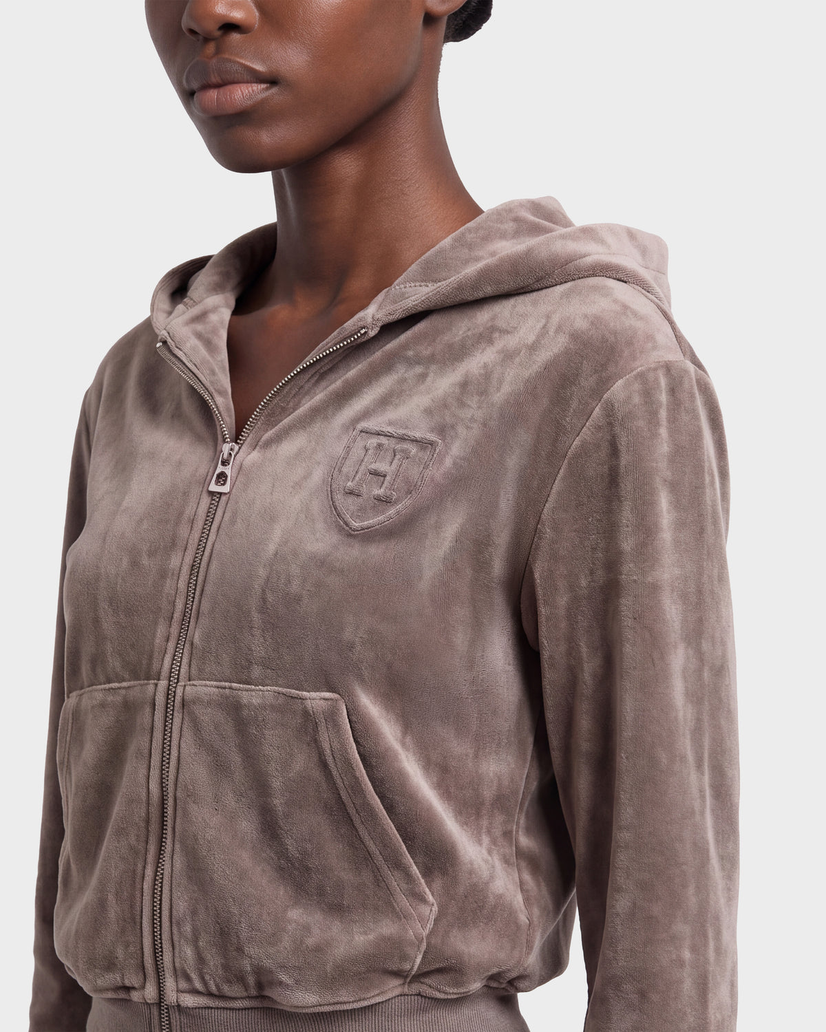 A person wearing the Rudsak TYRA HV - H526904 Taupe velour zip-up hoodie, featuring a front pocket and an embroidered emblem on the left chest, is shown from the shoulders to the waist against a plain background.