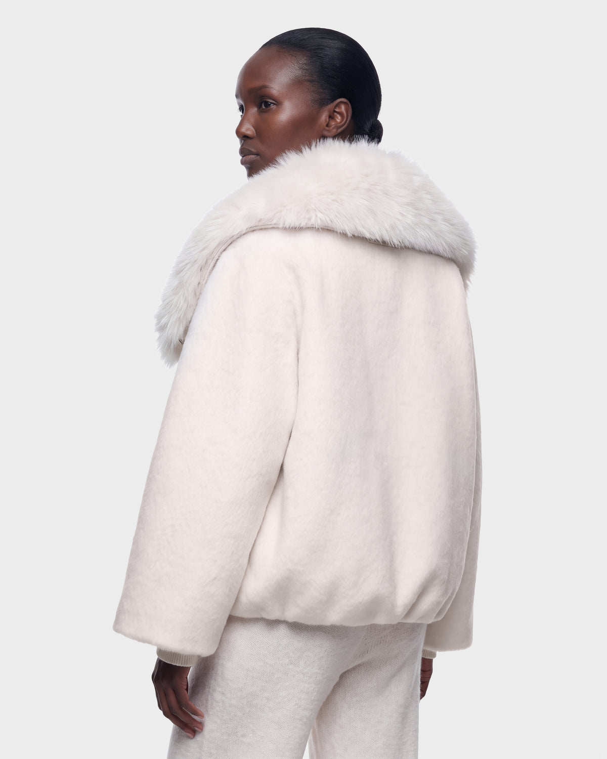 A person wearing the Rudsak ZARIA - 8126999 Vanilla light cream jacket with a large, fluffy fur collar and matching pants stands against a plain grey background, looking over their shoulder.