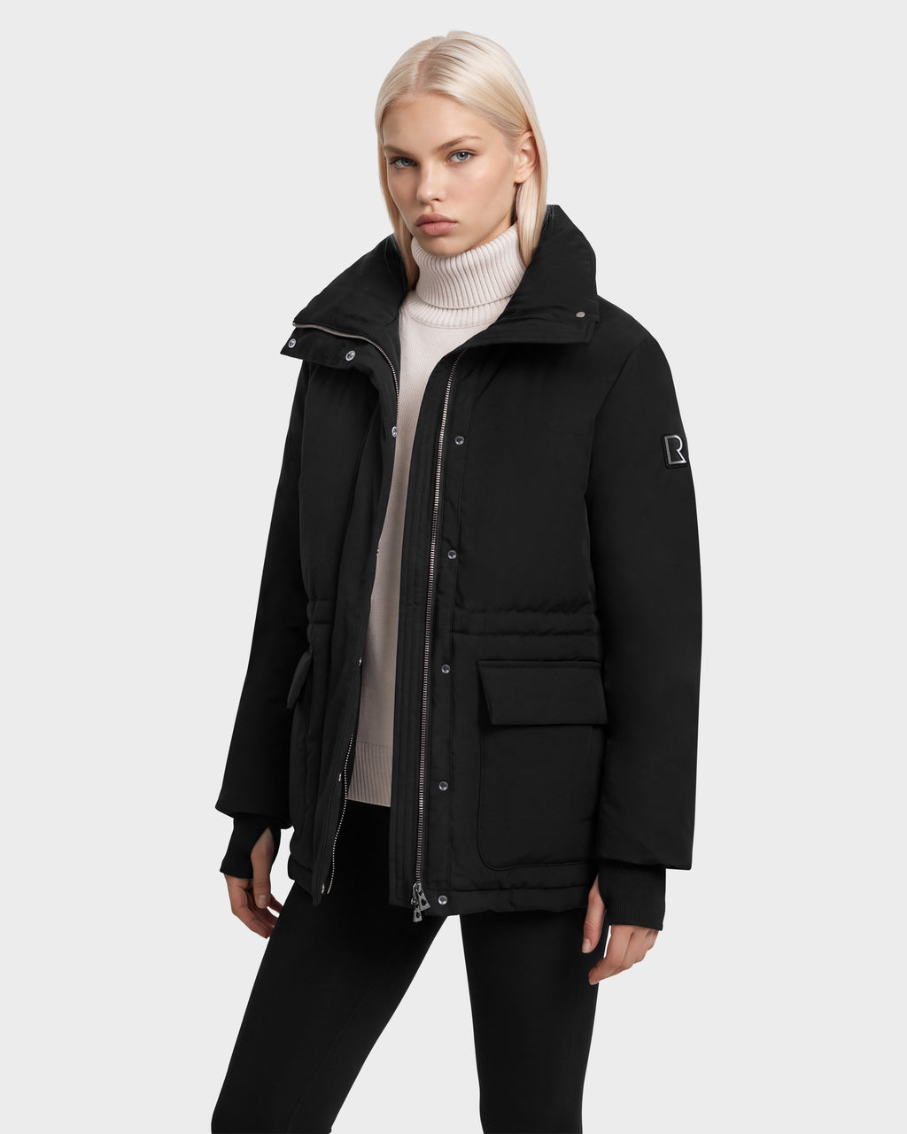 A person wears a black BHIMA - 8126515 mid-length parka by Rudsak with a high collar, front pockets, and a zippered closure over a cream turtleneck sweater.