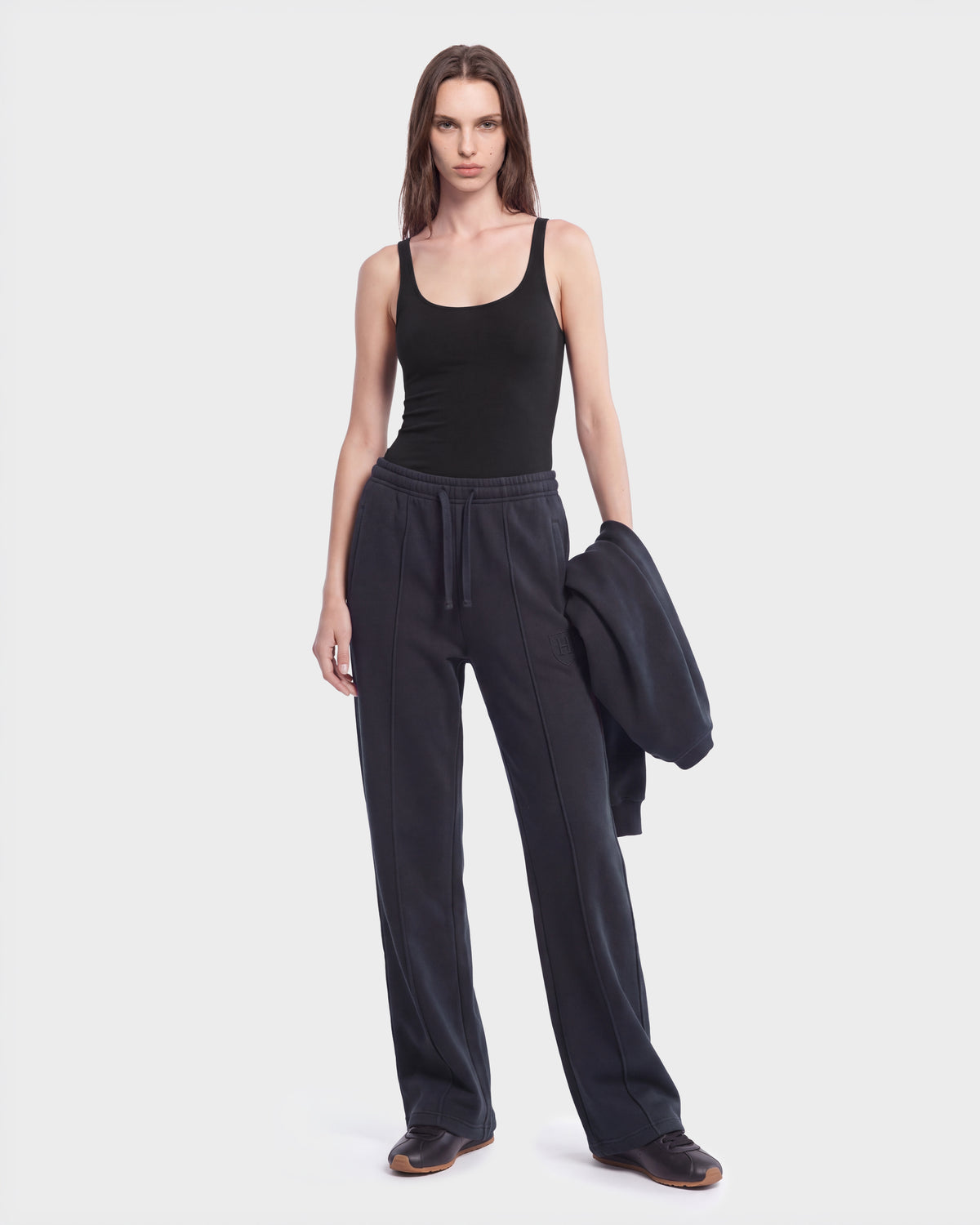 A person stands against a plain background wearing the Rudsak GAELLE HV - H526902 Titanium limited-edition heavyweight jersey joggers, paired with a black tank top and black shoes, holding a matching black jacket in one hand.