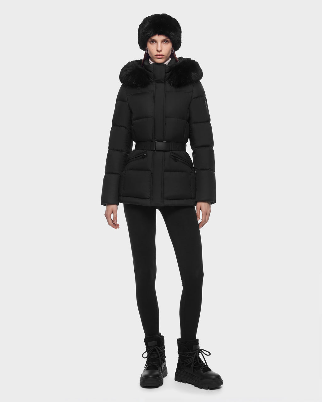 A model wears a Rudsak GRACE FF - 8126535 Black hooded puffer jacket with a belted waist, black leggings, a faux fur hat, and lug-sole boots.