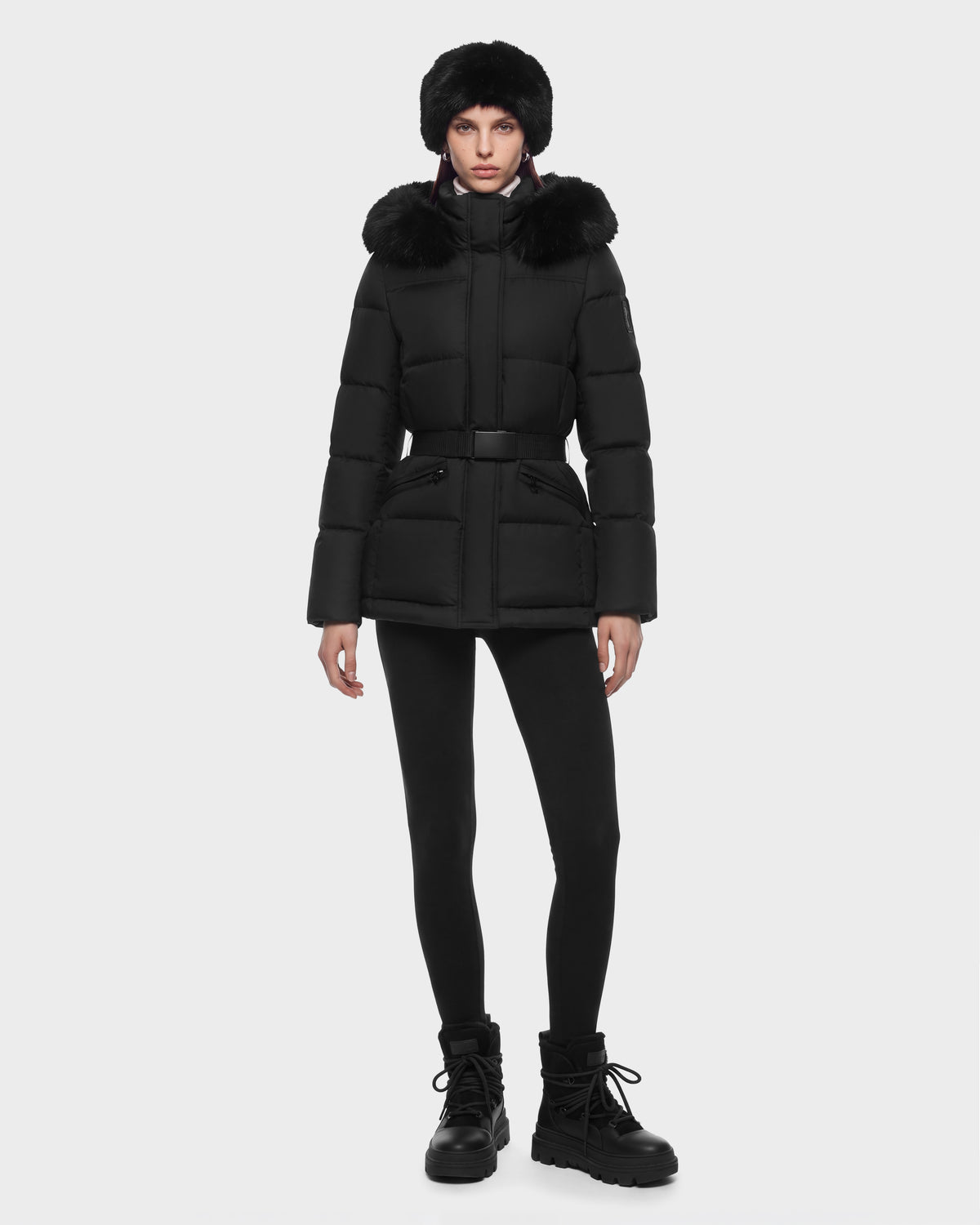 A model wears a Rudsak GRACE FF - 8126535 Black hooded puffer jacket with a belted waist, black leggings, a faux fur hat, and lug-sole boots.