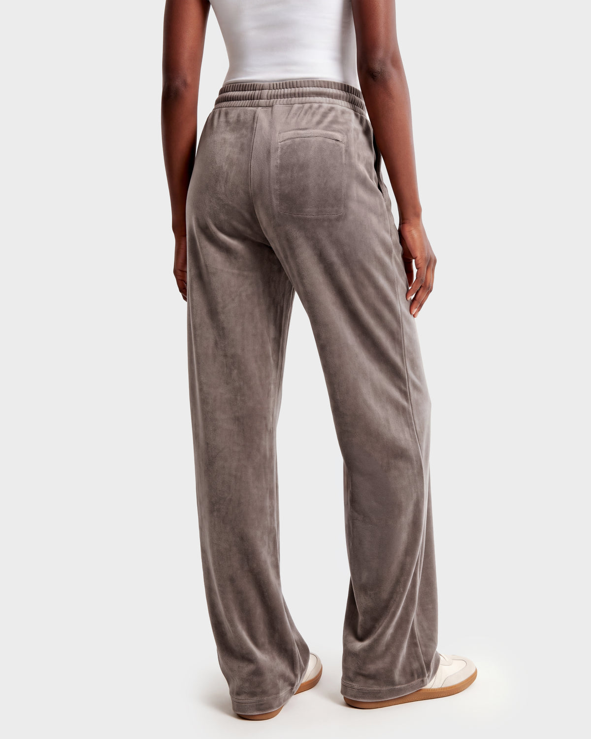 A person stands facing away, wearing the Rudsak OLIVYA HV - H526903 Taupe velour pants with a back pocket and elastic waistband, paired with a white tank top and casual white shoes. The plain, light background highlights the light brown taupe color of the pants.