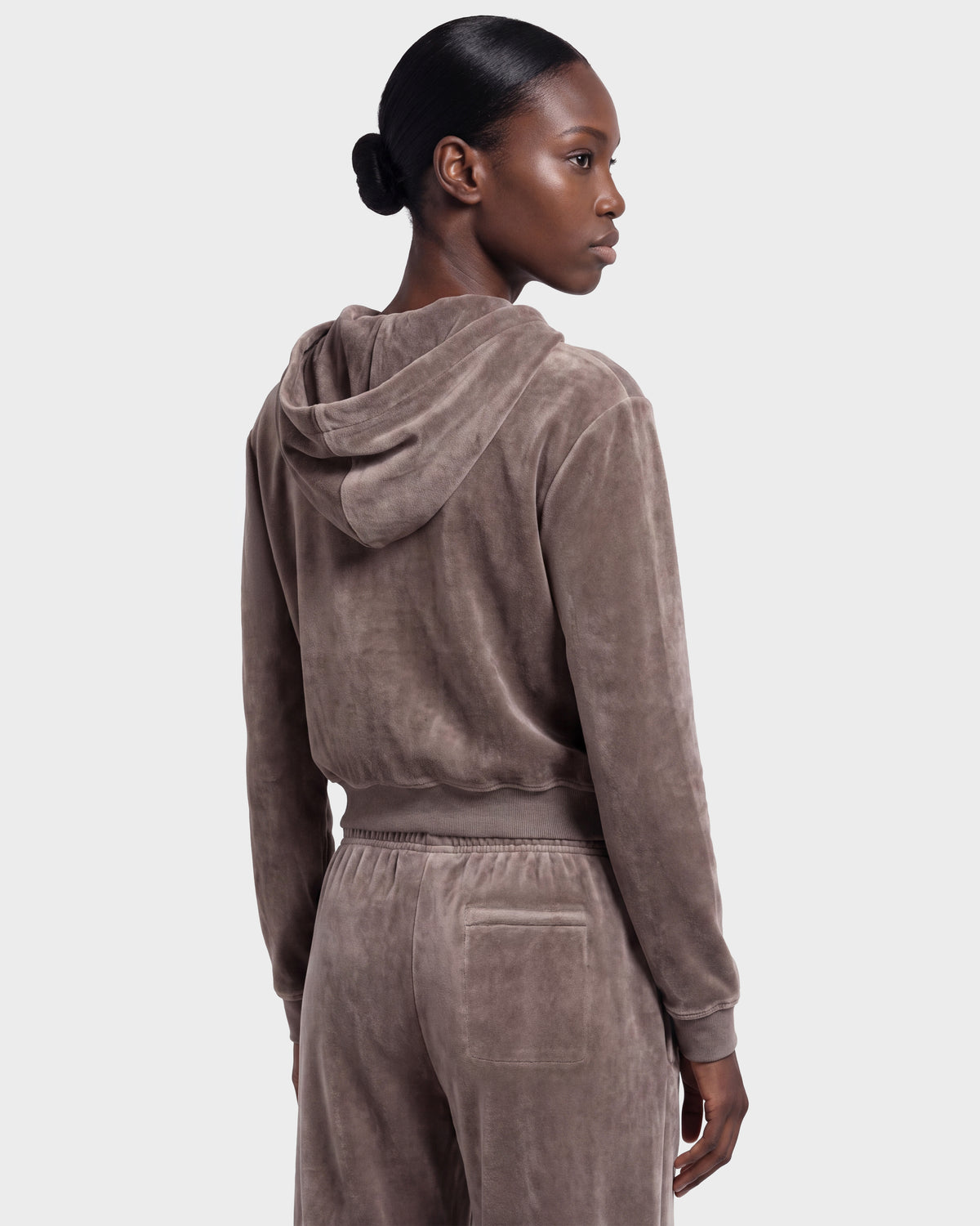 A woman with her hair in a low bun is seen from the back wearing the Rudsak TYRA HV - H526904 tracksuit in taupe, set against a plain light gray background.
