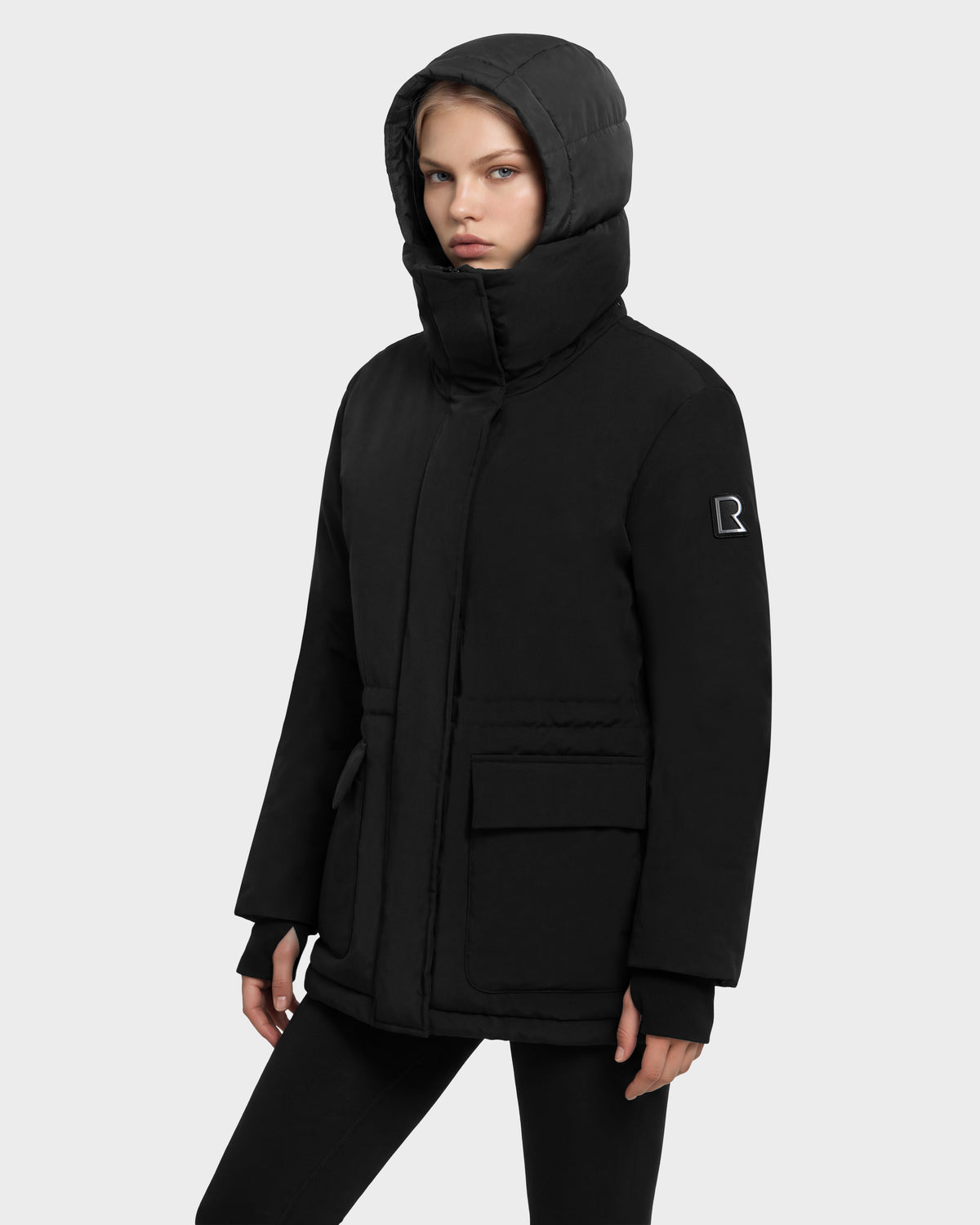 A model wearing a Rudsak BHIMA - 8126515 Black hooded parka with a high collar, two front flap pockets, and an emblem on the left sleeve.