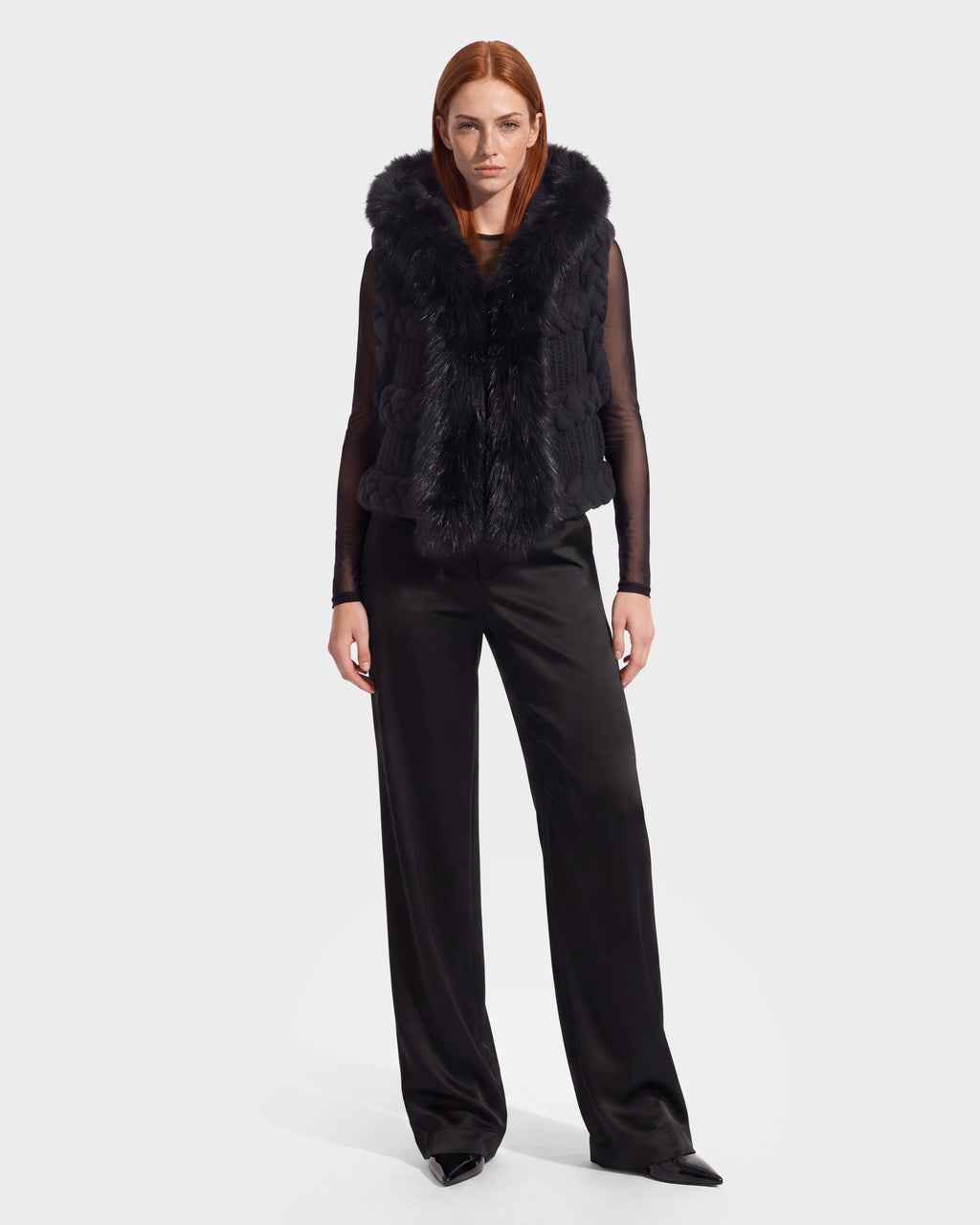 A person with straight red hair wears the CICELY - 8526686 Black fur vest by Rudsak—a nod to winter layering—over a sheer black top and loose black pants, standing against a plain light background.
