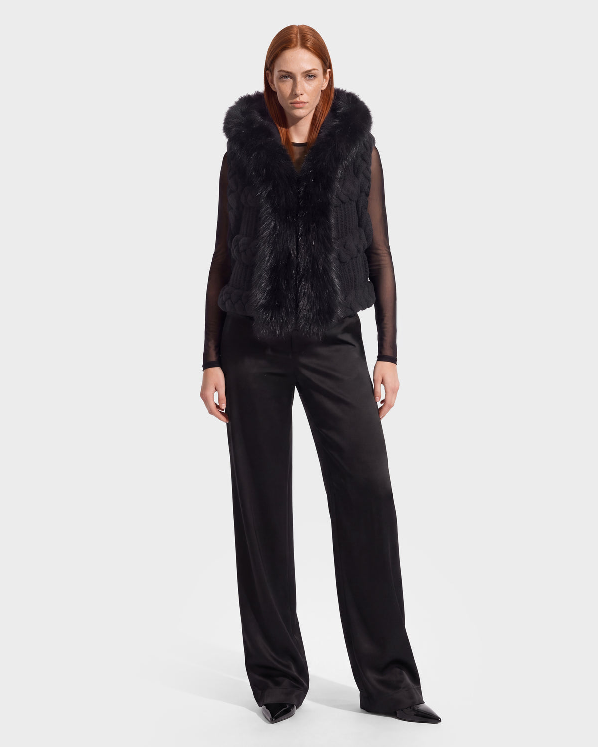 A person with straight red hair wears the CICELY - 8526686 Black fur vest by Rudsak—a nod to winter layering—over a sheer black top and loose black pants, standing against a plain light background.