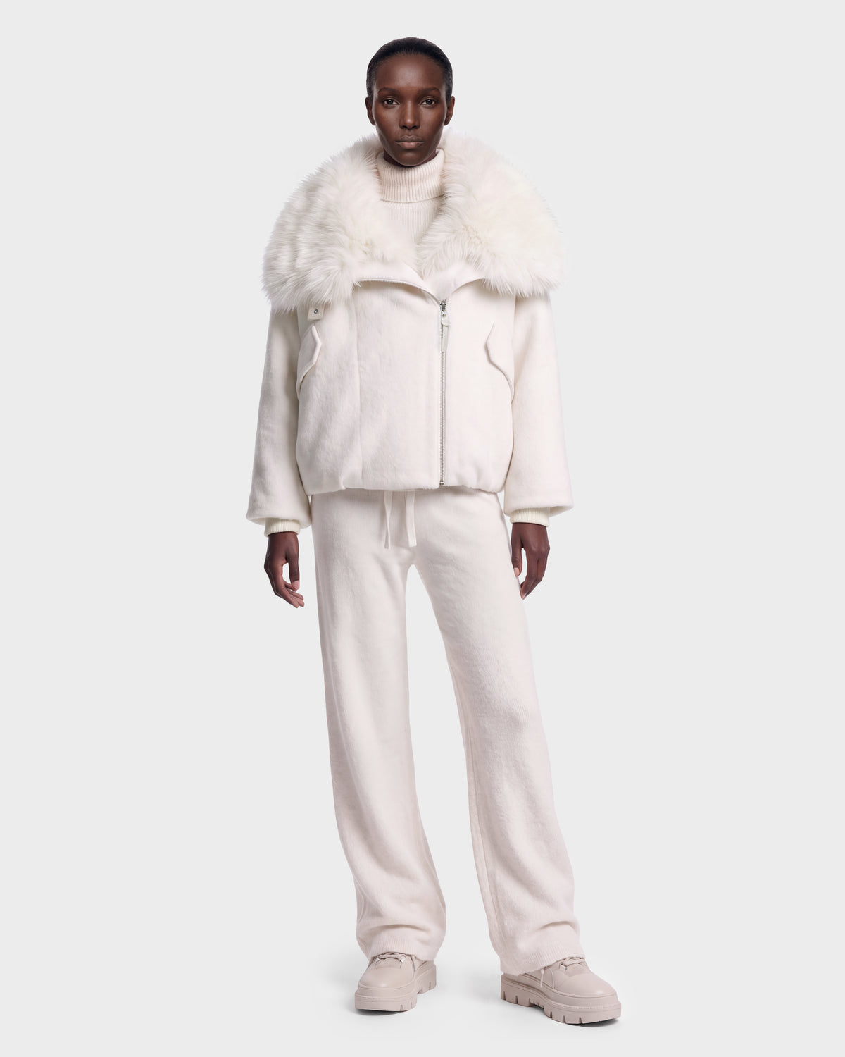 A person wearing the Rudsak ZARIA - 8126999 Vanilla faux fur-collared jacket, paired with a white turtleneck, loose pants, and chunky shoes, stands against a plain light background.
