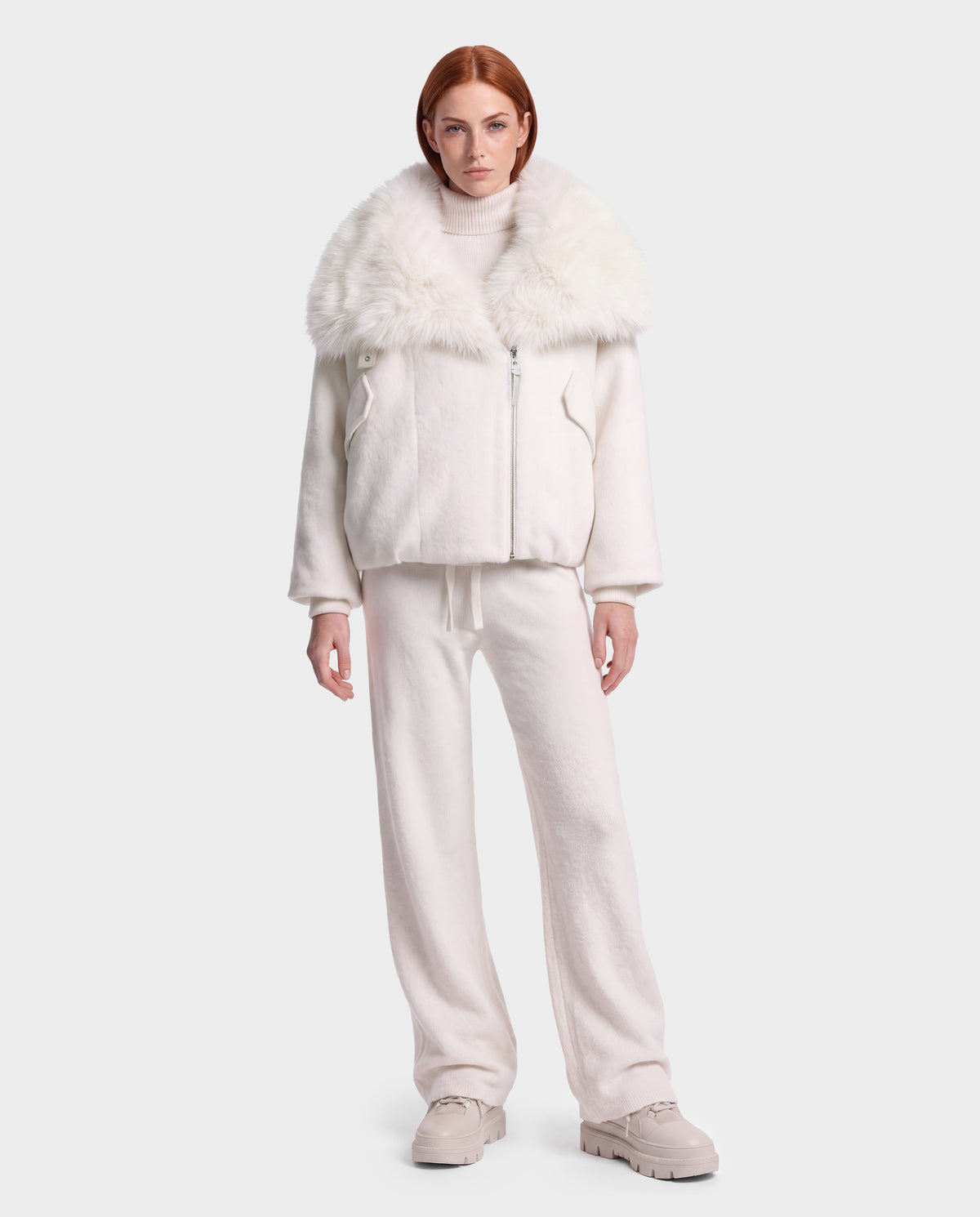 A woman wearing a Rudsak ZARIA - 8126999 Vanilla cream-colored winter puffer jacket with a large faux-fur collar, matching knit pants, and light-colored sneakers.