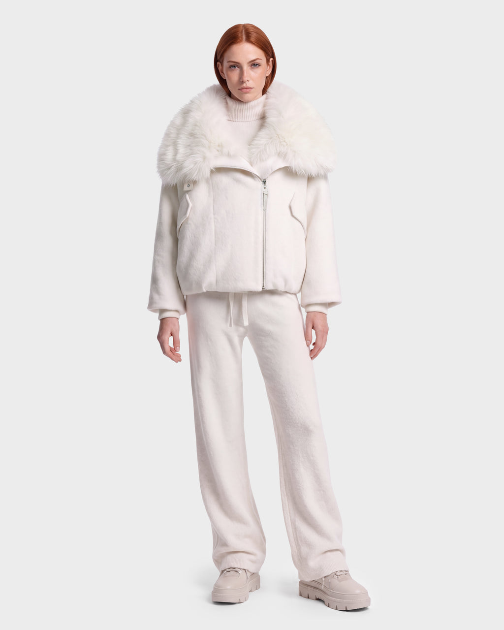 A woman with red hair stands against a plain light background, wearing the Rudsak ZARIA - 8126999 Vanilla oversized white winter puffer jacket with a large faux-fur collar, paired with a white turtleneck sweater, loose white pants, and beige shoes.