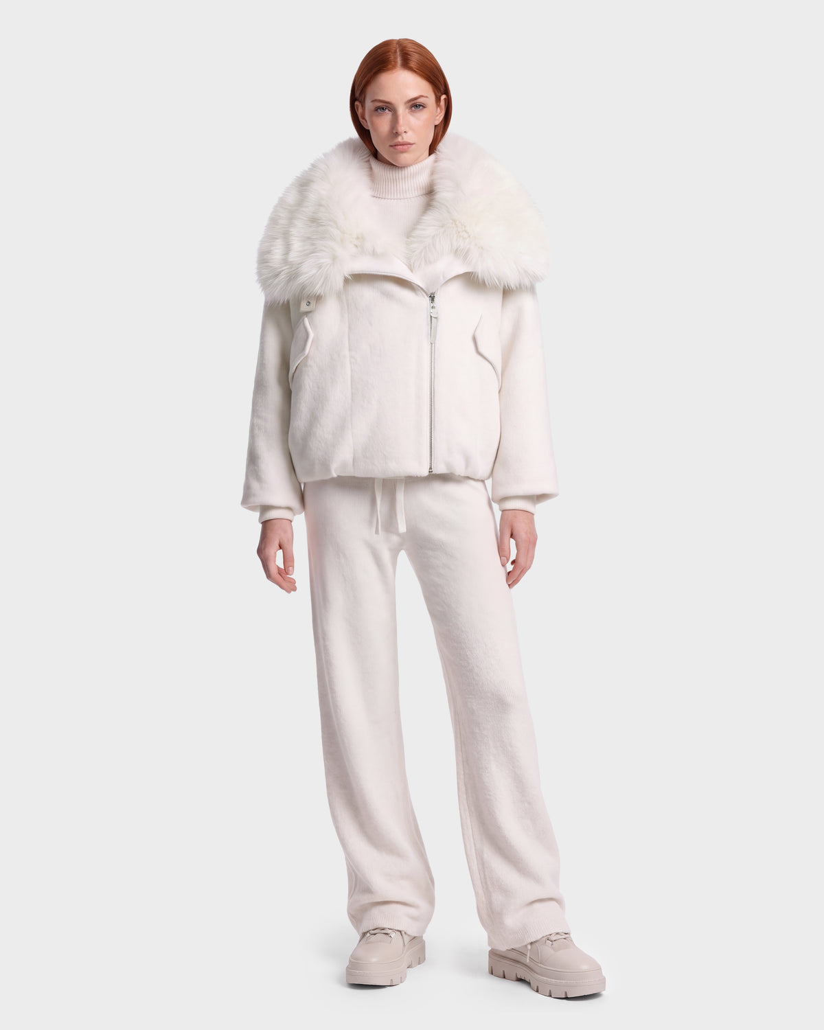 A woman with red hair stands against a plain light background, wearing the Rudsak ZARIA - 8126999 Vanilla oversized white winter puffer jacket with a large faux-fur collar, paired with a white turtleneck sweater, loose white pants, and beige shoes.