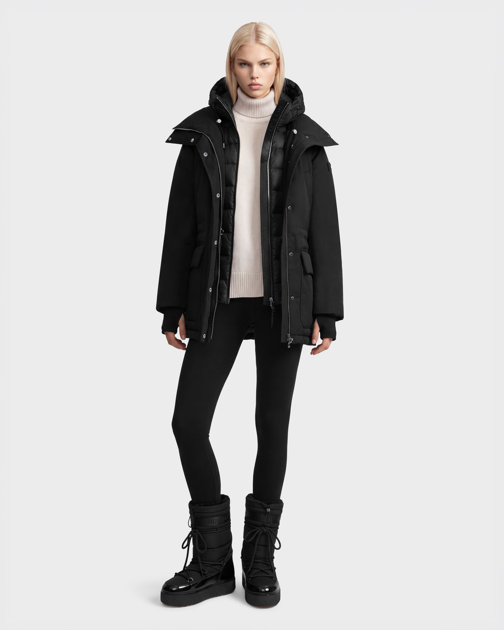 A person wears a black Rudsak BHIMA - 8126515 Black hooded winter parka over a beige turtleneck sweater, black leggings, and black winter boots.