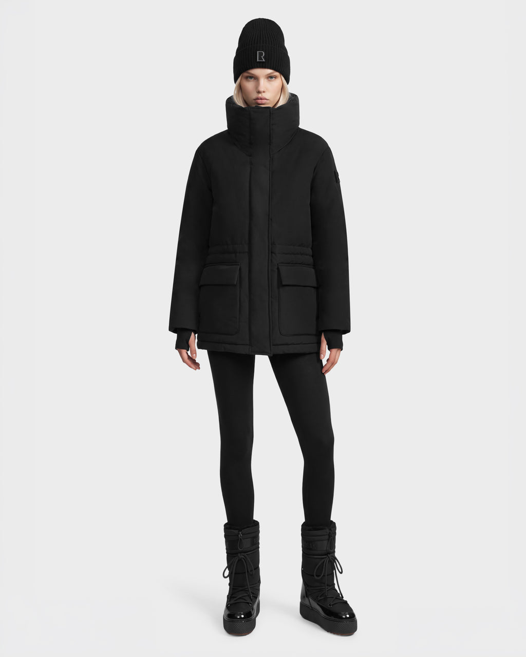 Model wearing a black BHIMA - 8126515 Black parka with a high collar, black leggings, a black beanie with a logo, and black lace-up winter boots.