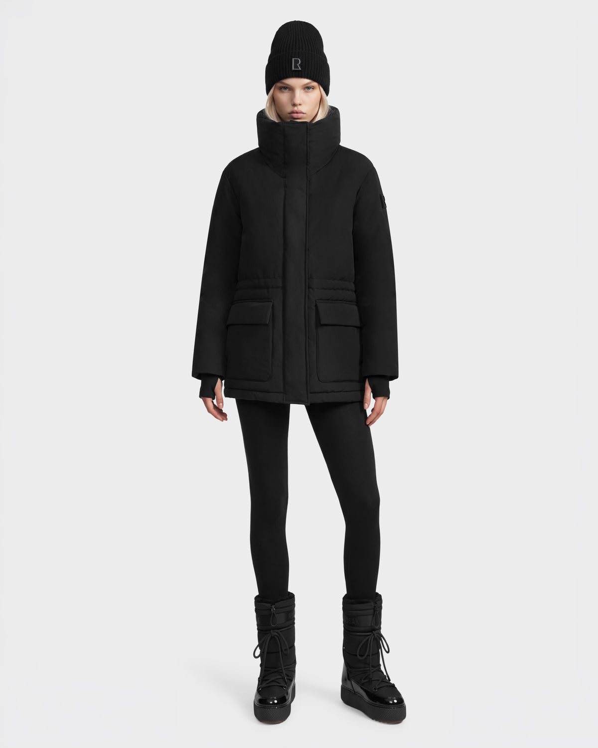 Model wearing a black BHIMA - 8126515 Black parka with a high collar, black leggings, a black beanie with a logo, and black lace-up winter boots.