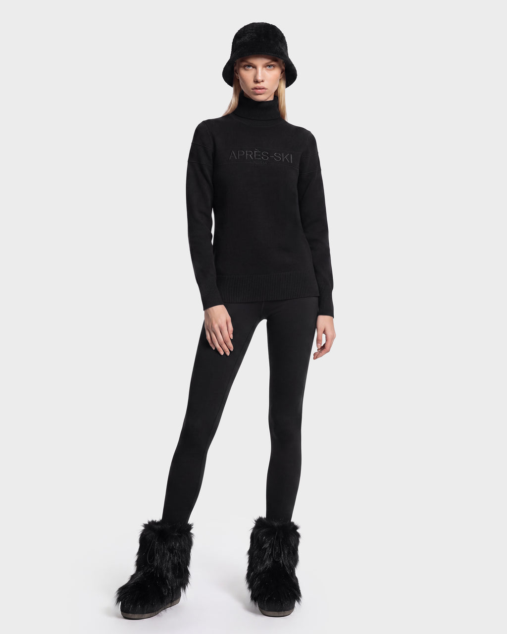 A person wearing the ERIKA - 8525705 Black turtleneck sweater by Rudsak, paired with a black bucket hat, black leggings, and black furry boots stands against a plain white background—a chic addition to any winter wardrobe.