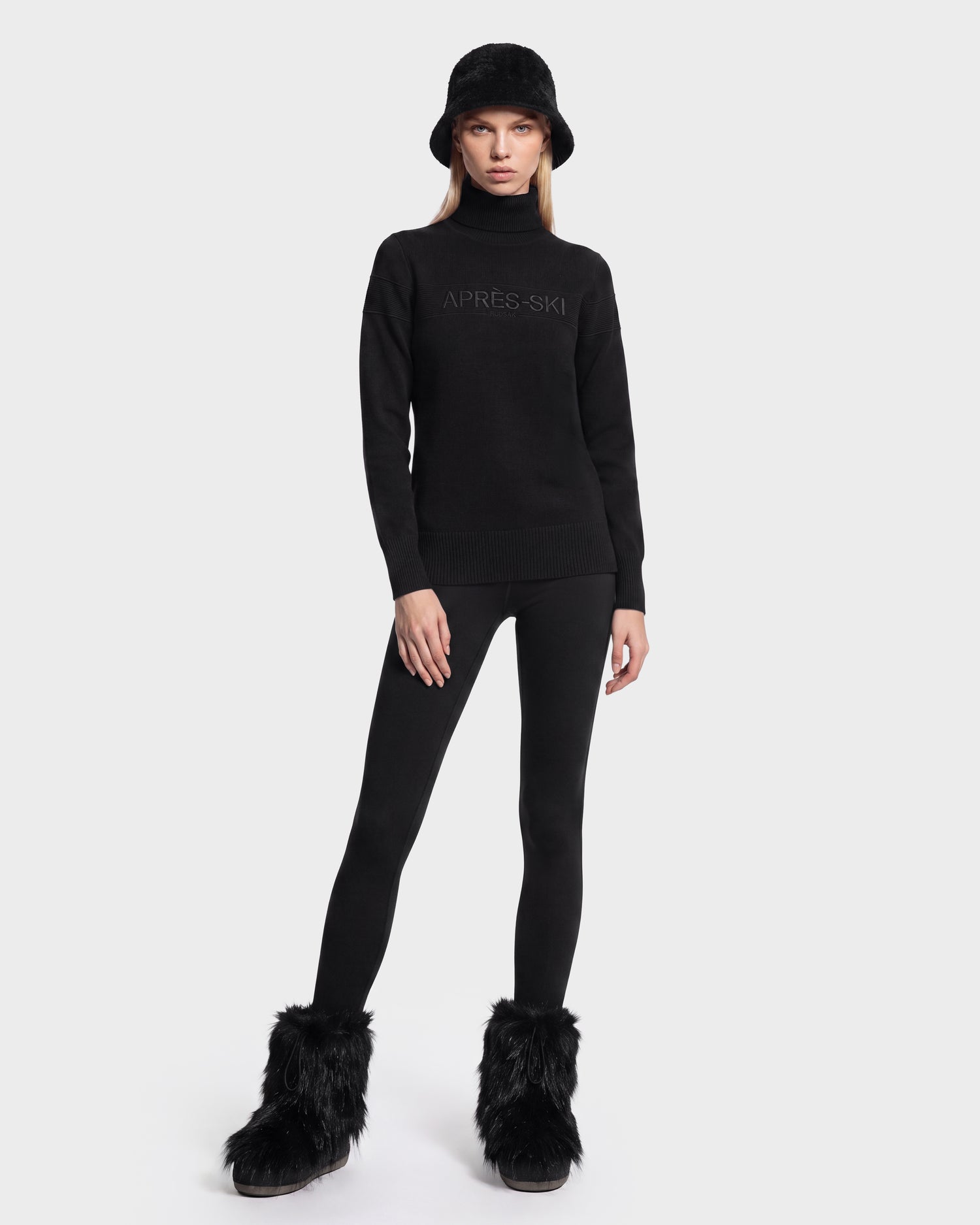 A person wearing the ERIKA - 8525705 Black turtleneck sweater by Rudsak, paired with a black bucket hat, black leggings, and black furry boots stands against a plain white background—a chic addition to any winter wardrobe.