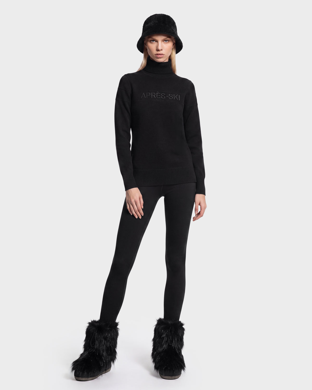 A person wearing the ERIKA - 8525705 Black turtleneck sweater by Rudsak, paired with a black bucket hat, black leggings, and black furry boots stands against a plain white background—a chic addition to any winter wardrobe.