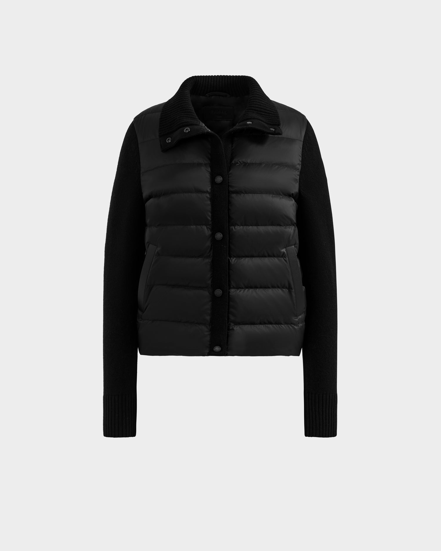 The ELLIE - 8125635 Black by Rudsak is a women’s quilted down puffer jacket with button-up front, ribbed cuffs, collar, sleeves, side pockets, and a padded body. Shown against a plain light gray background.