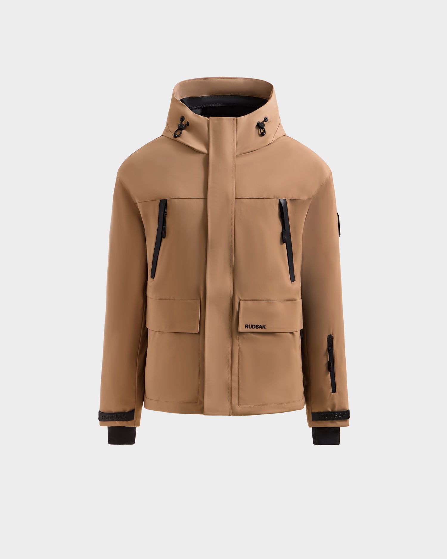 The ETHAN - 6125589 Sand by Rudsak is a tan men’s ski jacket with a hood, black zippers, front flap pockets, and adjustable cuffs, shown on a plain light background. Its modern minimalist design is ideal for winter sports.