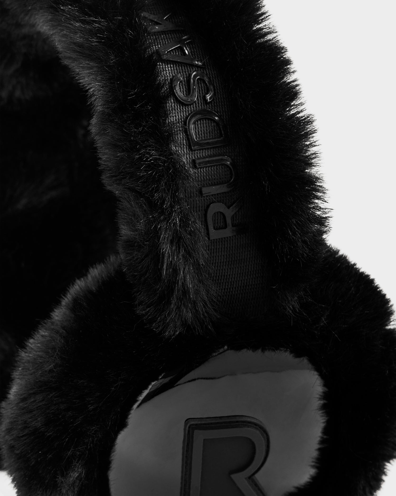 Close-up of the black VALENCIA - 8425543 Rudsak earmuff, featuring soft faux fur, an embossed Rudsak name on the band, and a raised R logo on the ear cover, set against a plain background.