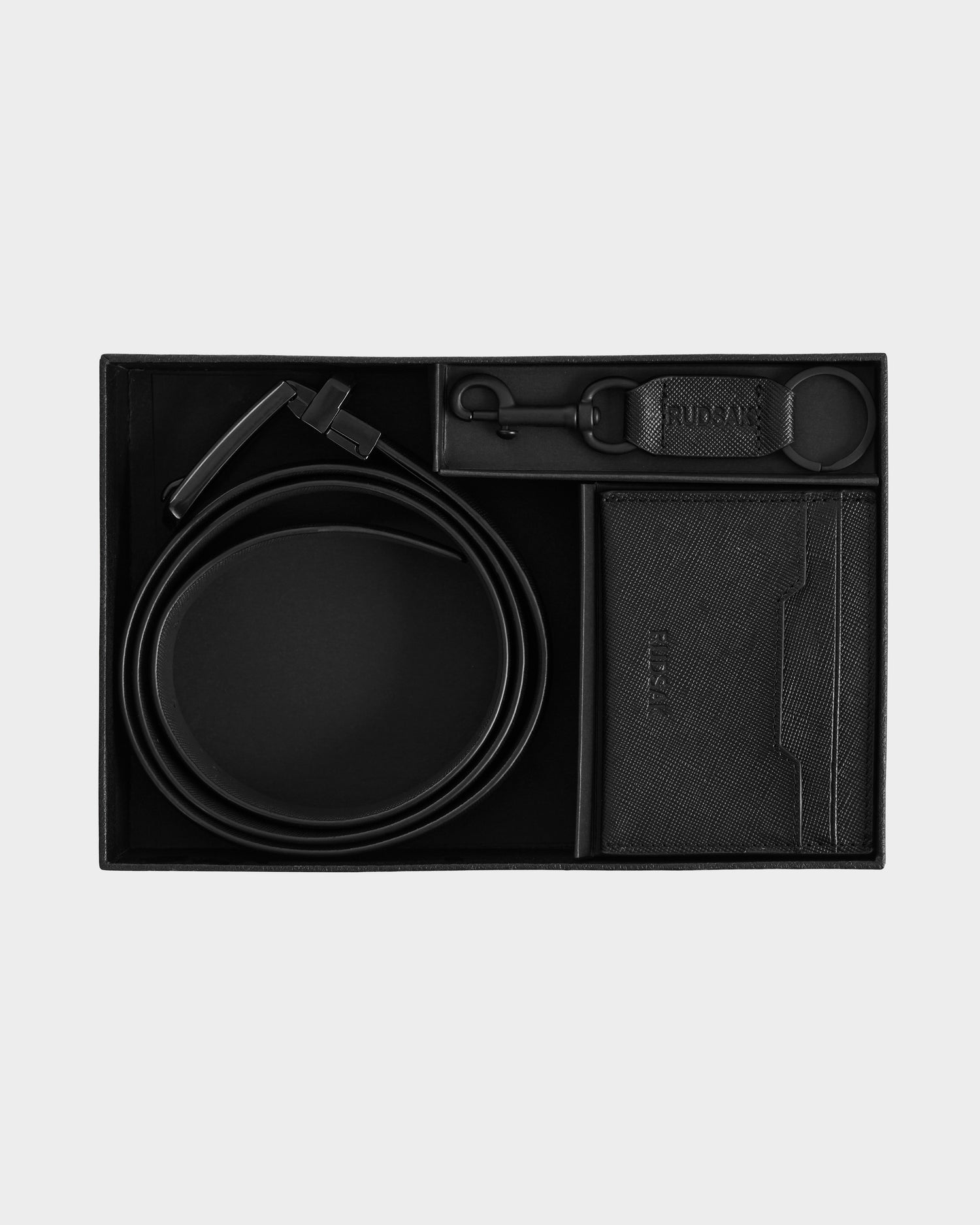 The Rudsak RYLIE - M424500 Black men’s box set includes a reversible leather belt, keychain, and cardholder, all stylishly presented in a black gift box against a light gray background.