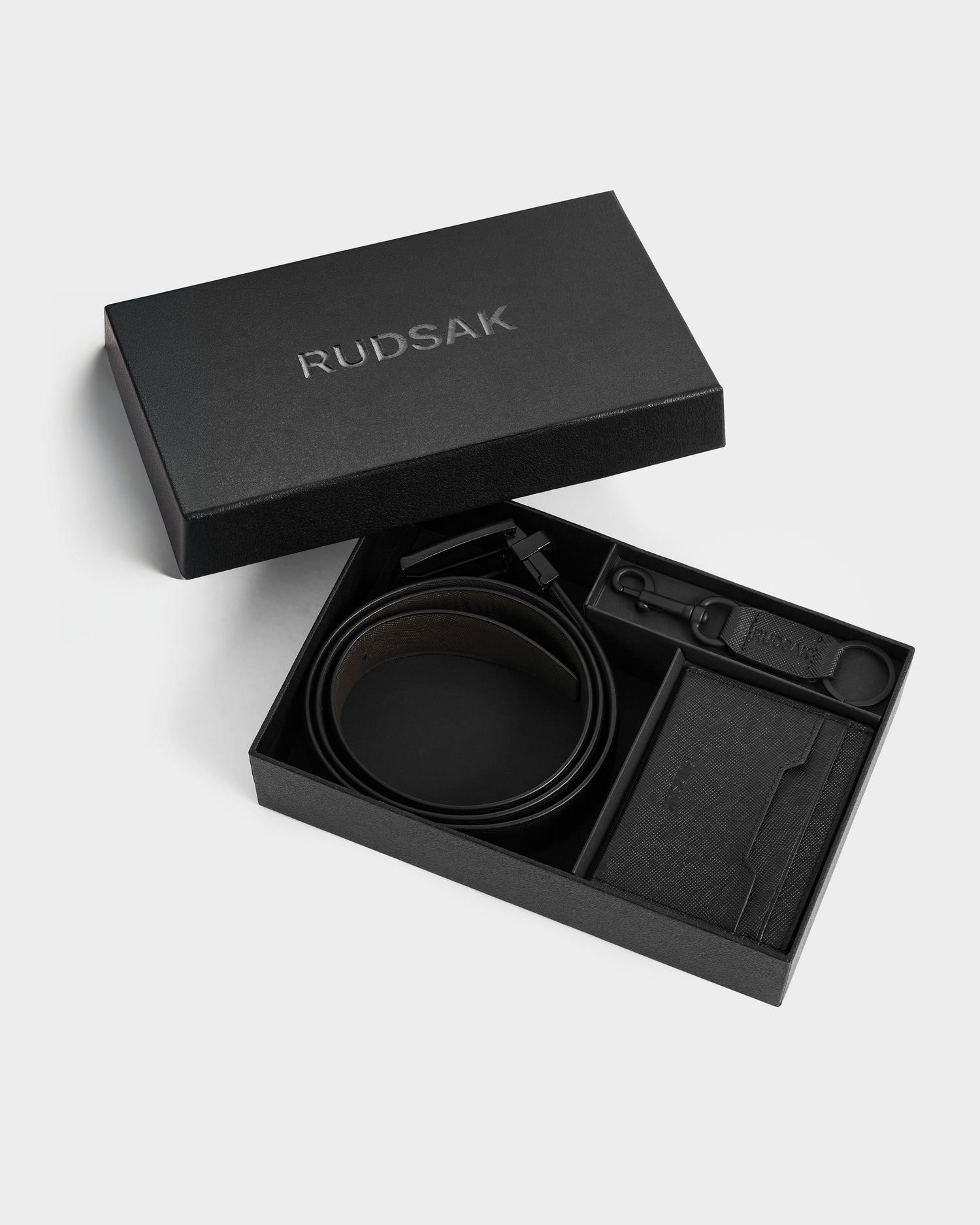 The RYLIE - M424500 Black by Rudsak features a reversible leather belt, cardholder, keychain, and small accessory arranged in a black Rudsak gift box with separate compartments.