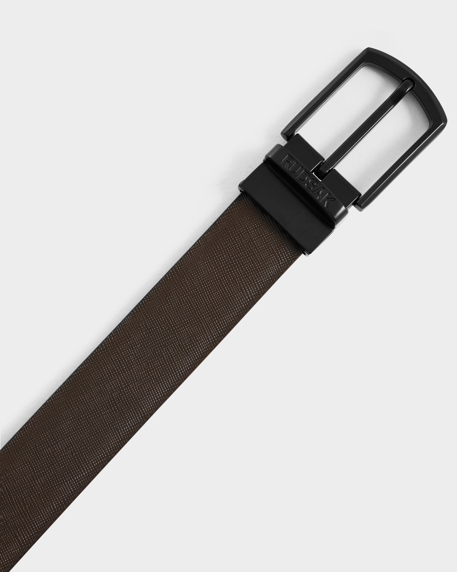 A close-up of the Rudsak RYLIE - M424500 Black reversible leather belt, featuring brown textured leather and a black metal buckle, photographed diagonally on a plain light background.