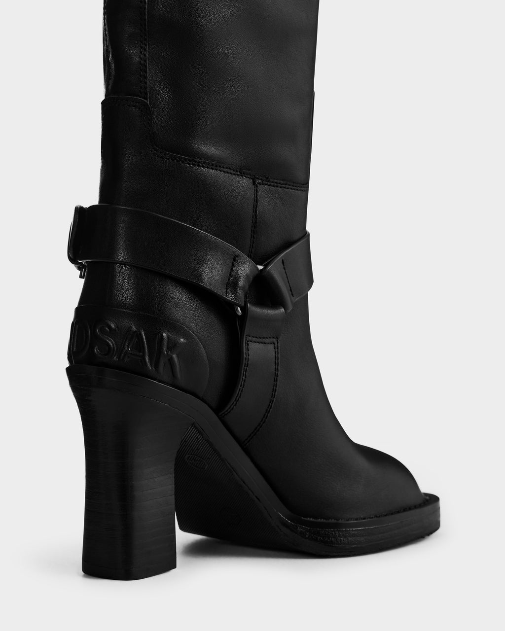A close-up of the ROSSEY - 8225574 Black boot by Rudsak, featuring a thick heel, smooth texture, decorative ankle strap, embossed logo near the back heel, and visible stitching.