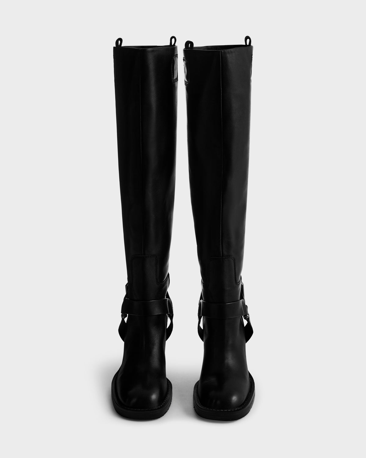 The Rudsak ROSSEY - 8225574 Black tall leather boots feature decorative straps and top loops, shown facing forward on a plain light gray background.
