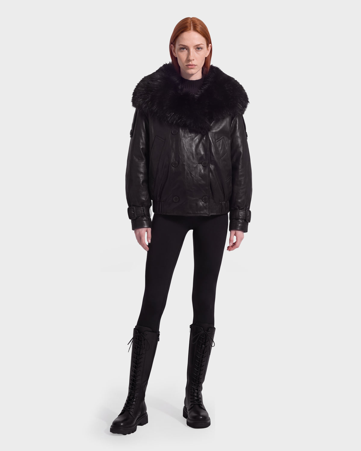 A person wearing the Rudsak JANELLE - 8126936 Black leather bomber with a large faux fur-trimmed collar, black leggings, and tall lace-up boots.