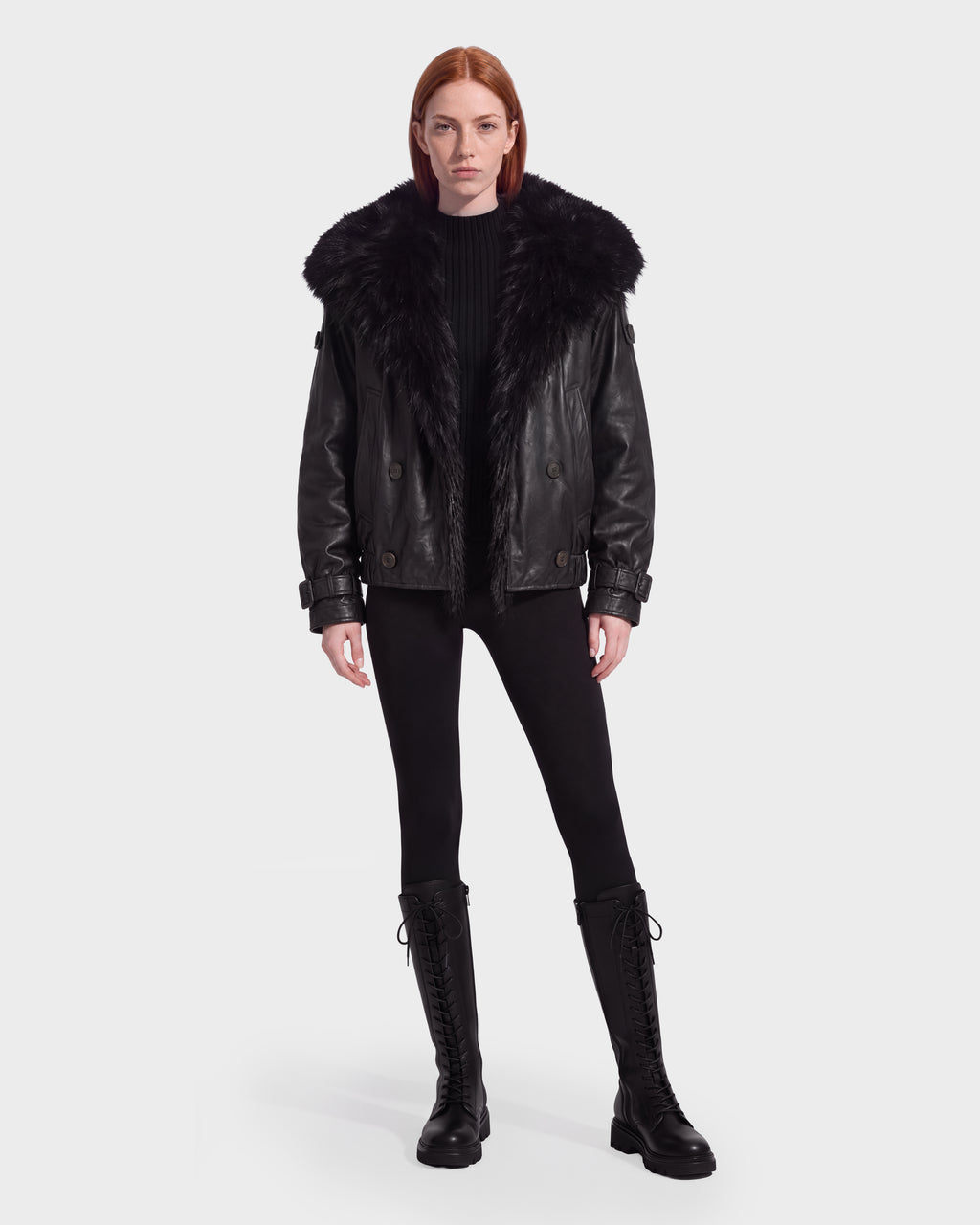 A woman wearing a Rudsak JANELLE - 8126936 Black leather bomber with a large faux fur collar over a black ribbed sweater, black leggings, and black lace-up knee-high boots.