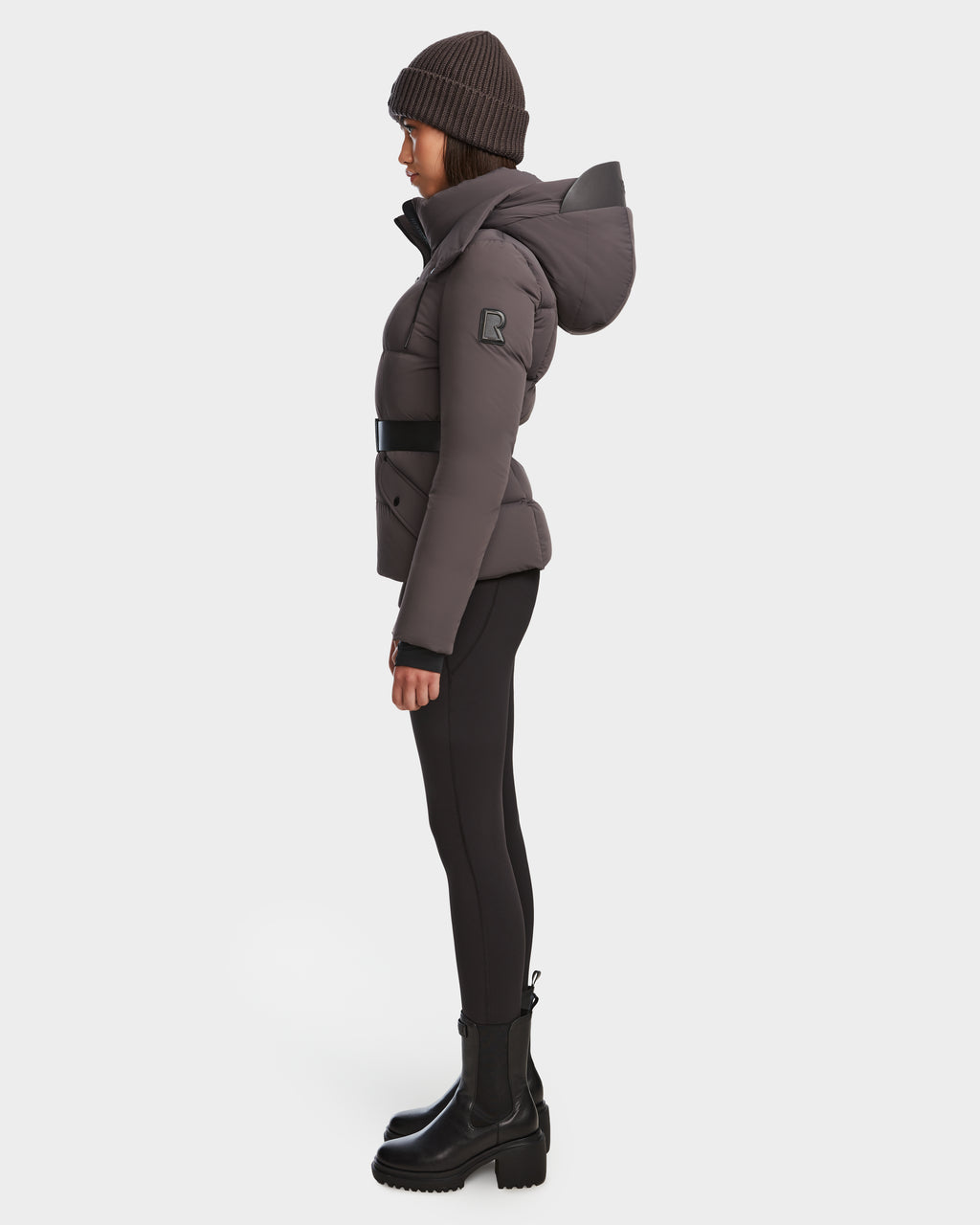 A person in side profile wears the Rudsak ADELINE - 8124544 Grey womens down puffer coat with hood and black belt, paired with black leggings, boots, and knit beanie, against a white background. The jacket features RDS-certified down insulation for warmth.