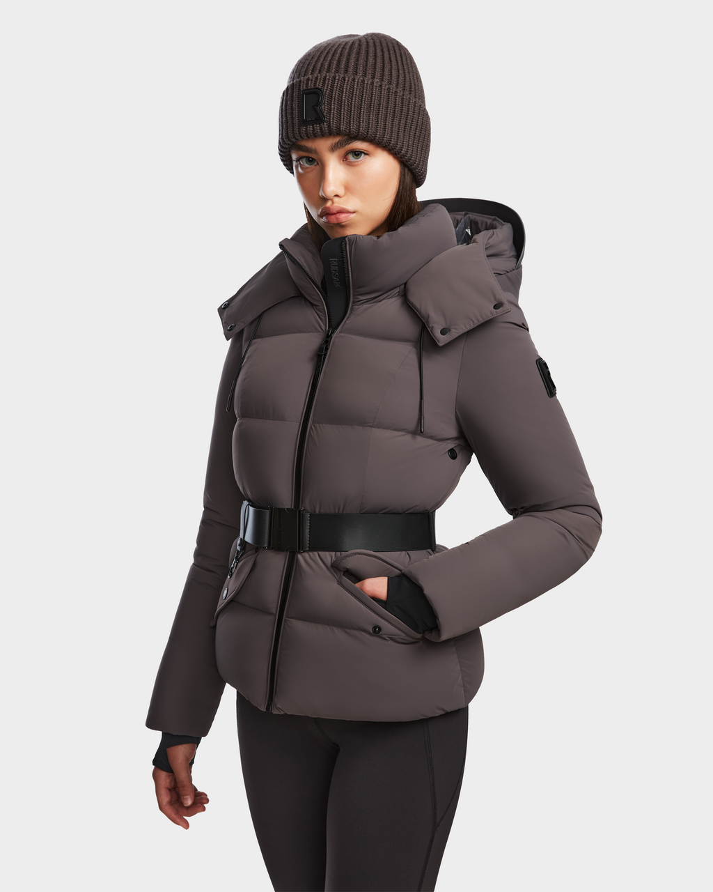 A woman wearing the Rudsak ADELINE - 8124544 Grey women’s down puffer with a black belt, black pants, and a brown knit beanie stands against a plain white background.