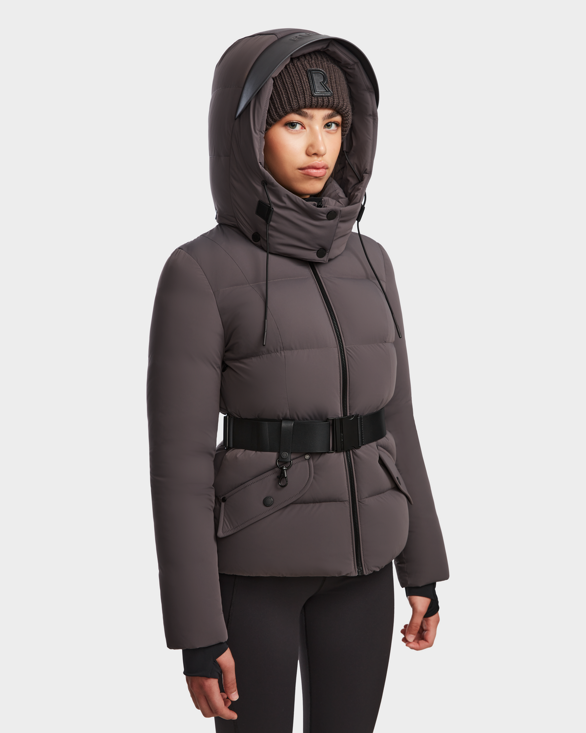 A woman wearing the Rudsak ADELINE - 8124544 Grey down puffer jacket with a belted waist, high collar, and hood, plus a black knit beanie and leggings, stands against a plain white background.