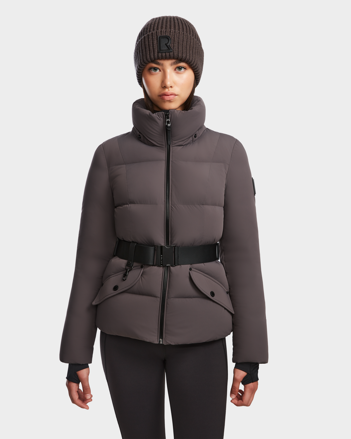 A woman wearing the Rudsak ADELINE - 8124544 Grey RDS-certified down puffer jacket with a black buckle belt, black leggings, and a dark brown knit beanie stands facing forward against a plain white background.