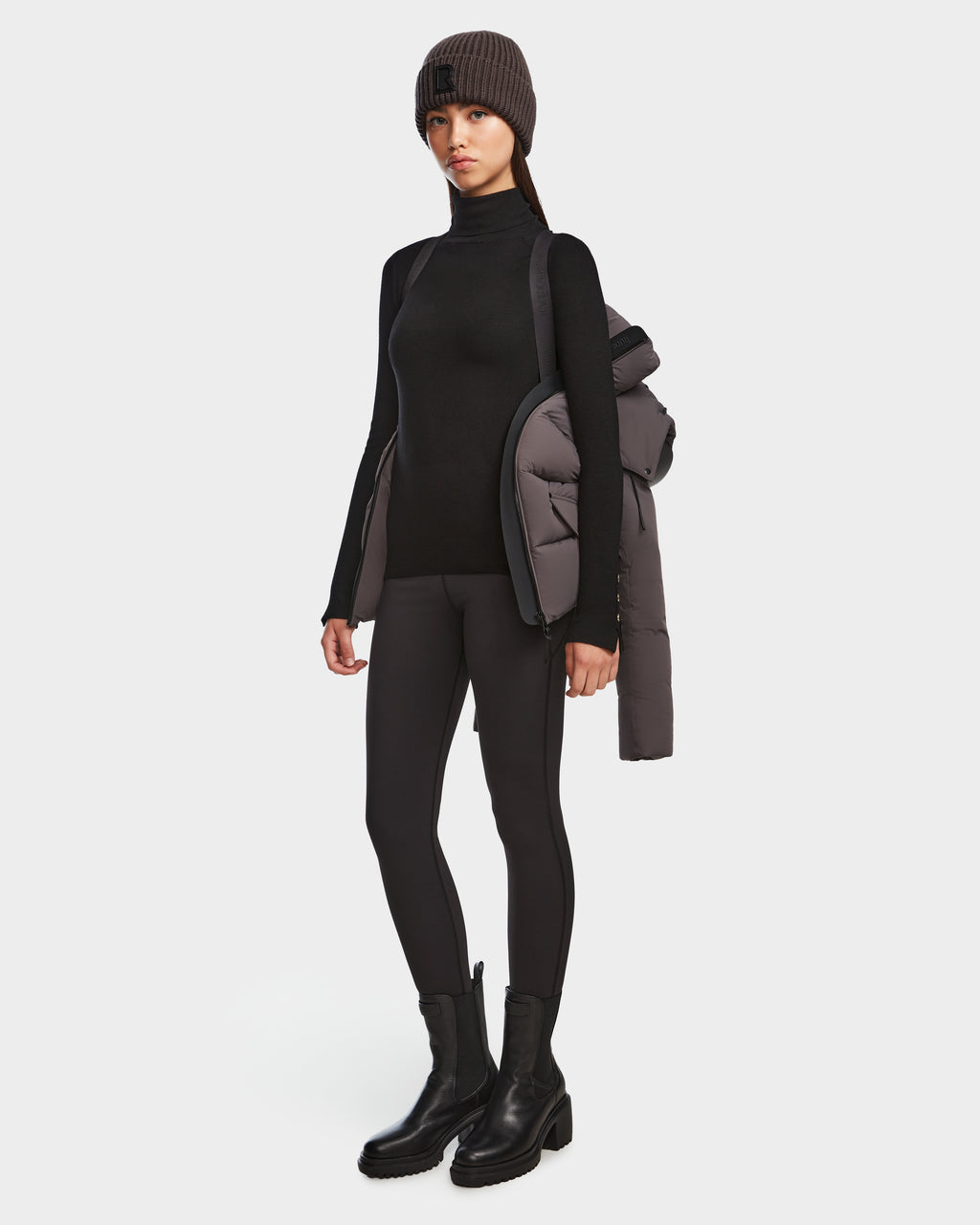 A woman poses against a white background in a black turtleneck, leggings, boots, and gray knit beanie, with the Rudsak ADELINE - 8124544 Grey RDS-certified down winter jacket draped over one shoulder.