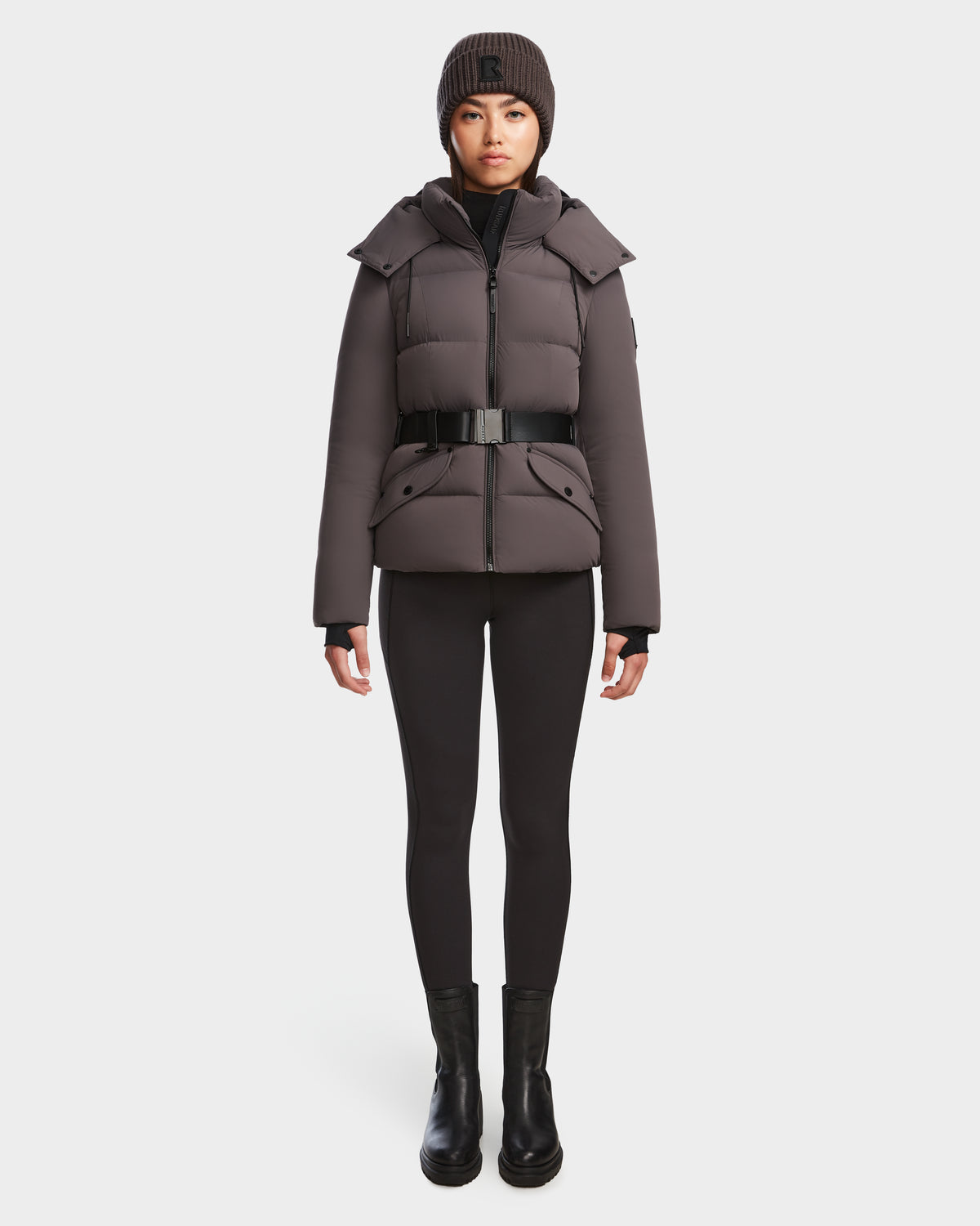 A person faces forward wearing the Rudsak ADELINE - 8124544 Grey womens down puffer jacket with a belt, paired with black leggings, black boots, and a dark knit beanie against a plain white background.
