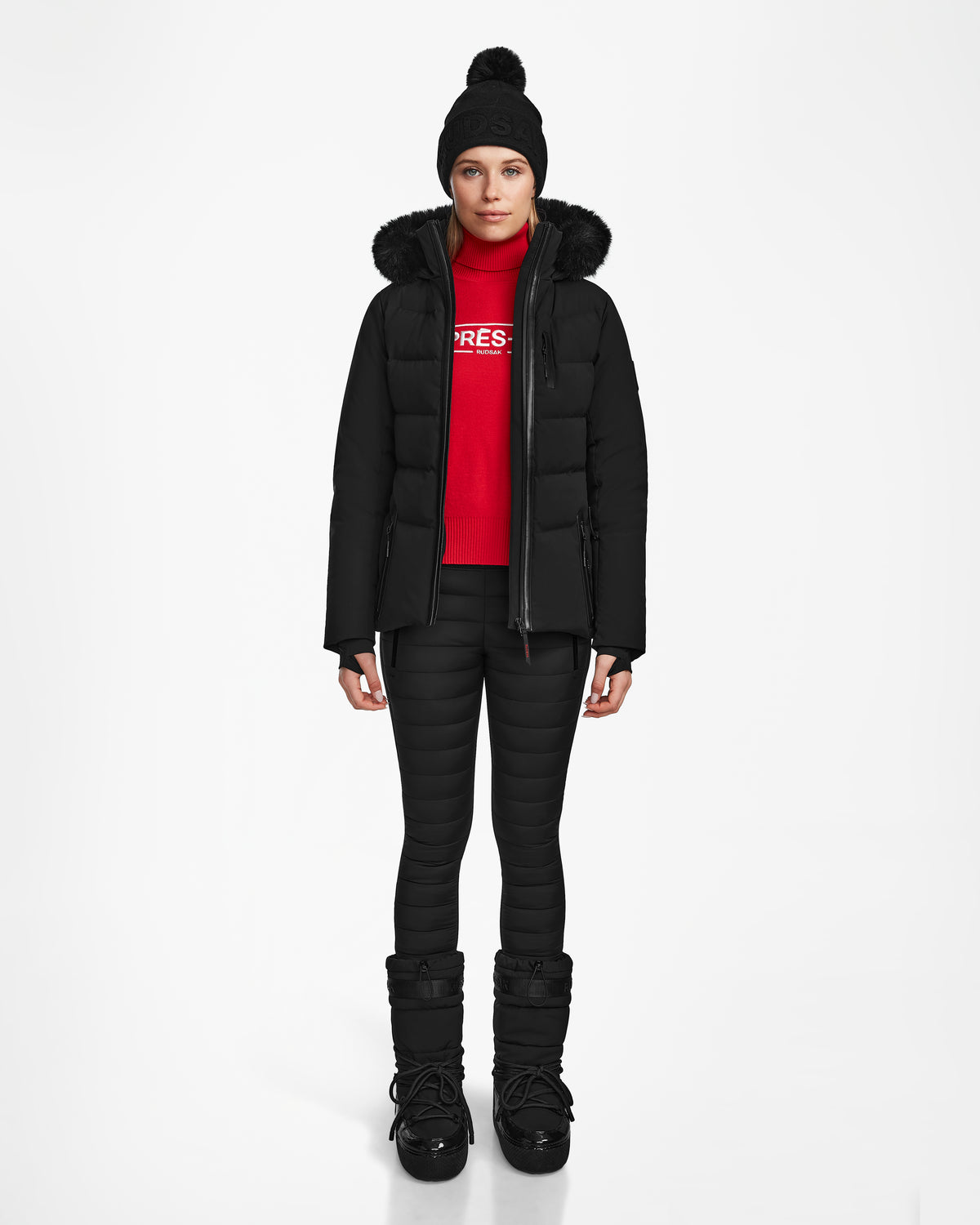 A person faces forward against a white background, wearing Rudsak’s ALAIA - 8124816 Black women’s quilted ski jacket with RECCO rescue technology, a black winter hat, red sweater, black quilted pants, and black winter boots.