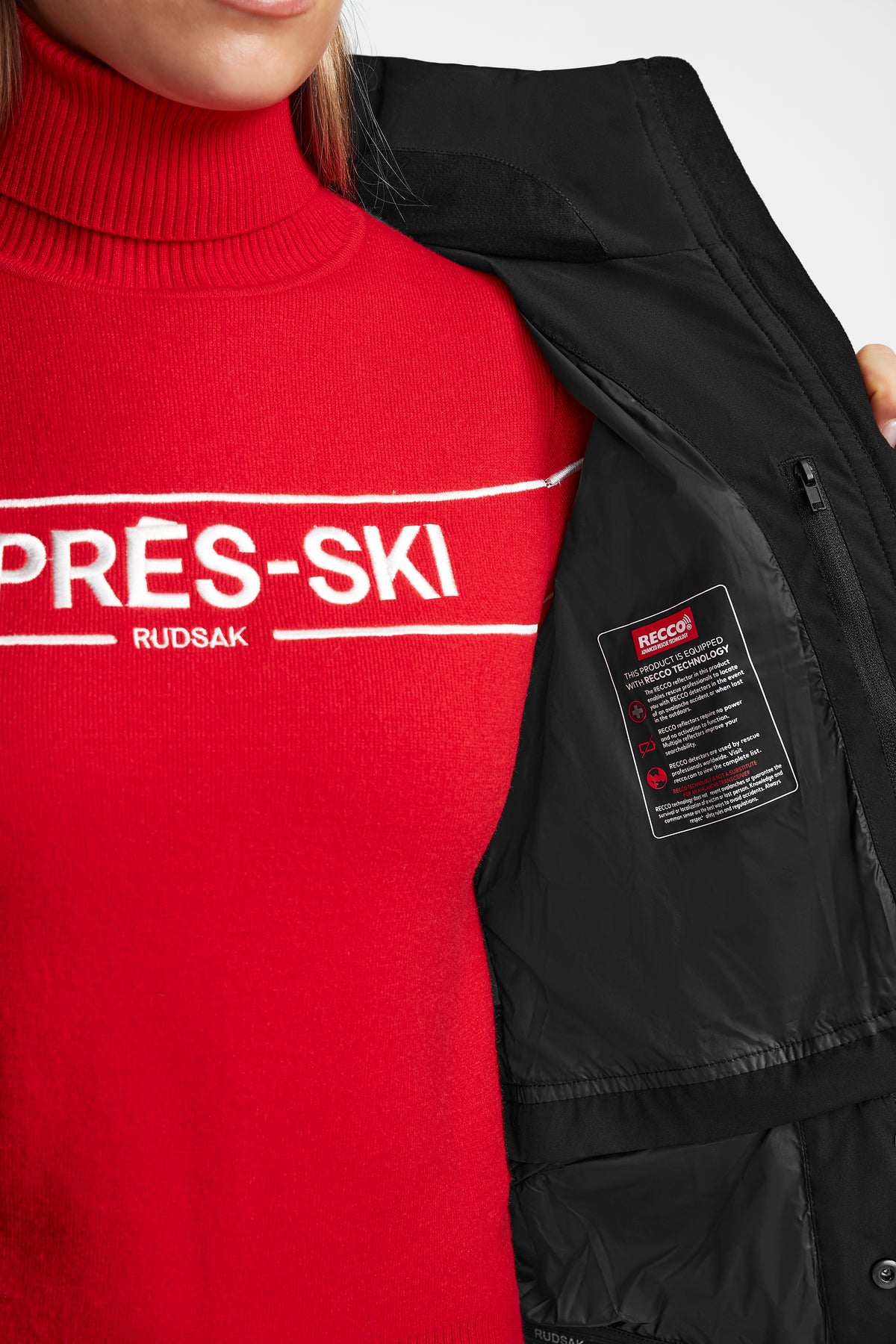 A person in a red turtleneck opens a Rudsak ALAIA - 8124816 Black women’s quilted ski jacket, revealing an interior label with ski-specific features and a RECCO rescue technology tag.